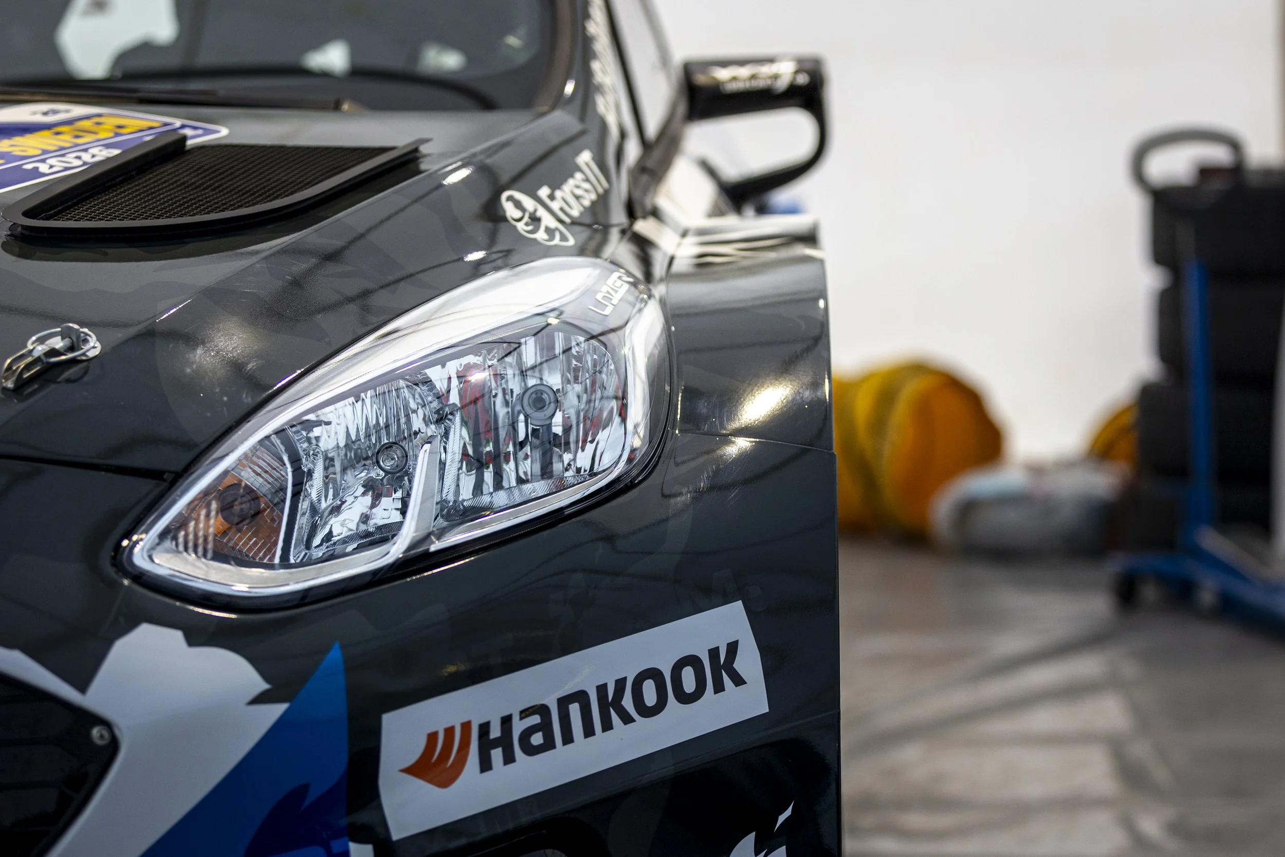 Close-up of a rally car with sponsor stickers, including Hankook, showing parts of the front headlight, hood, and a large rear wing, positioned indoors with racing equipment in the background.