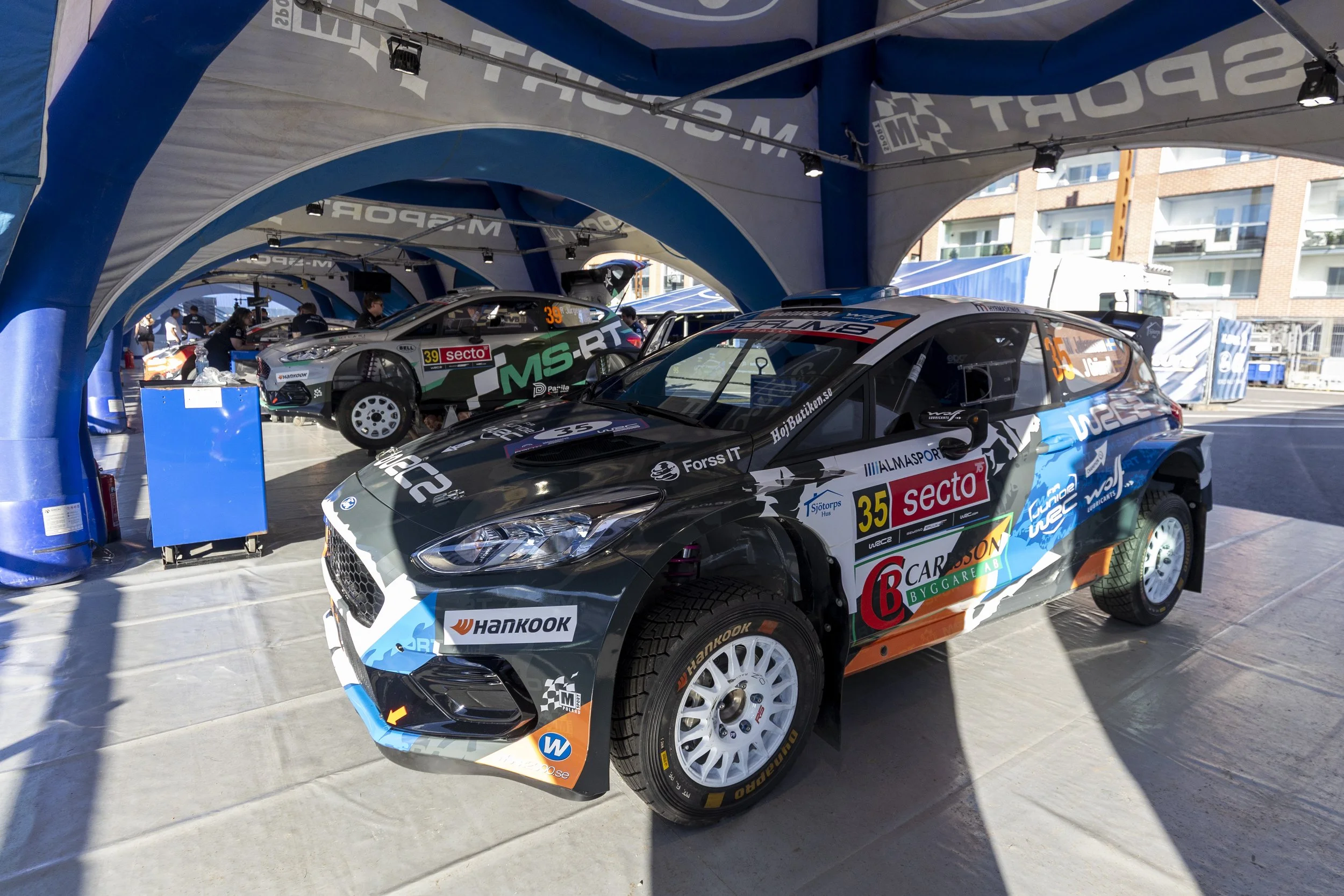 Race cars in a pit area under a large canopy, with team members working on the vehicles and various sponsor logos visible on the cars and canopy.