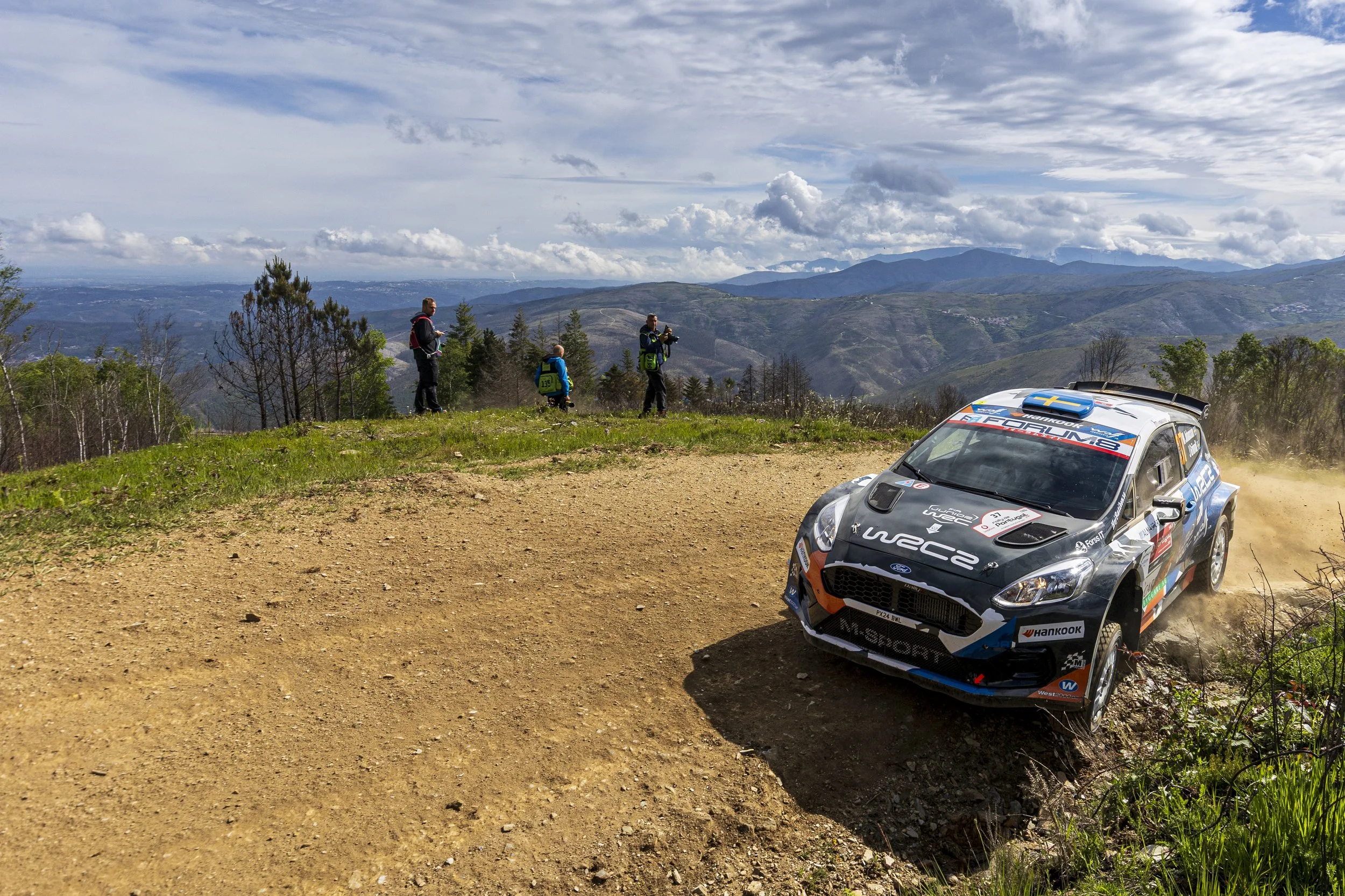 A rally car racing on a dirt track with a mountainous landscape in the background, and spectators standing on the side of the track.