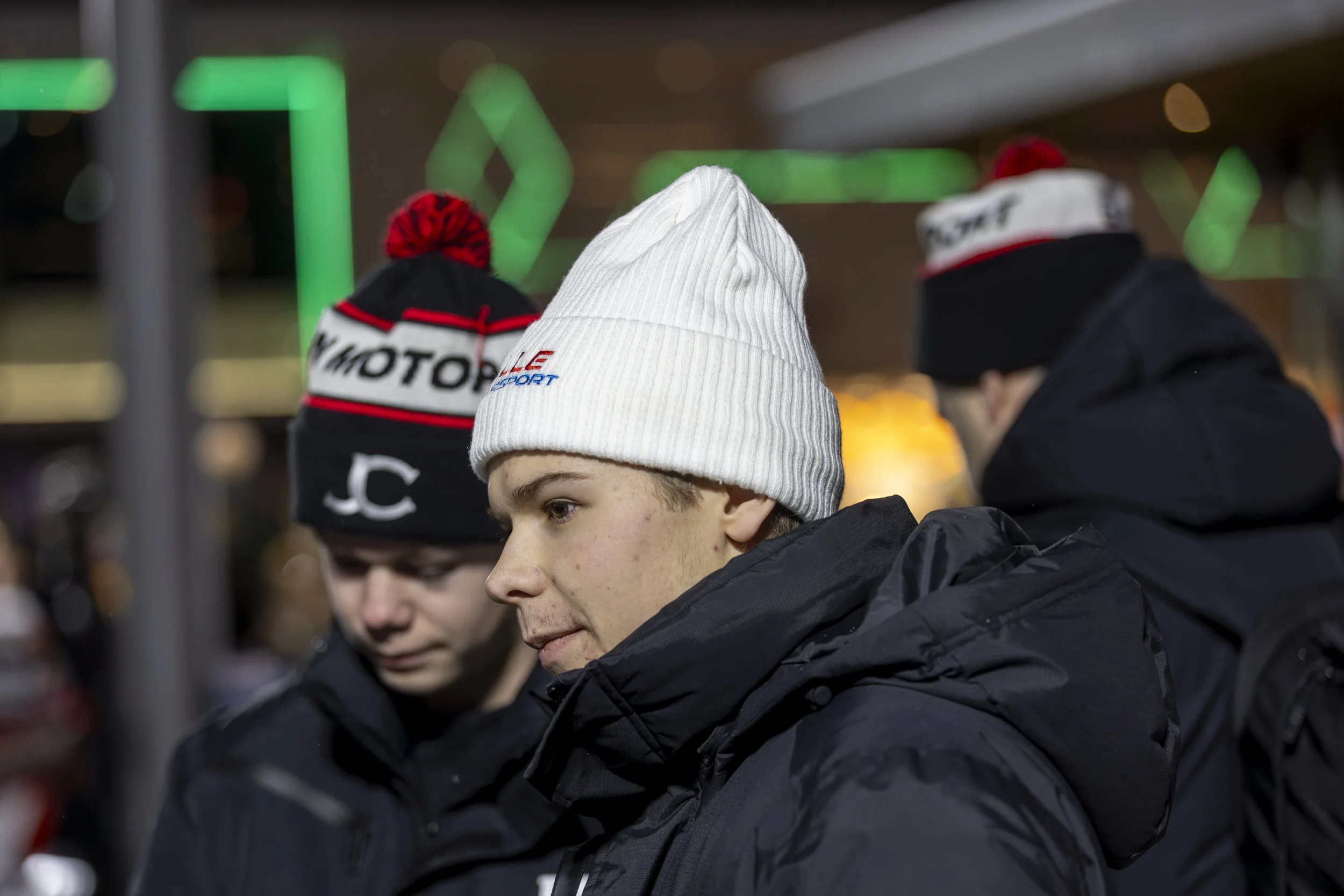 Close-up of two young men wearing Beanie hats, one white and one black, at a nighttime outdoor setting with green lights in the background.