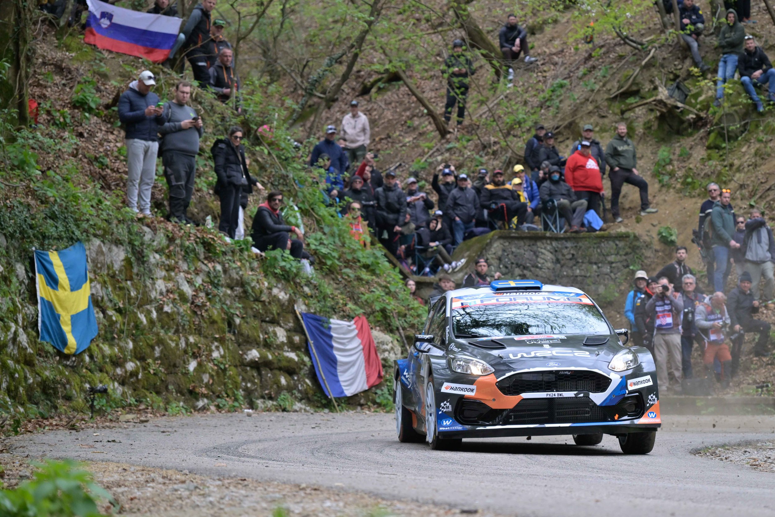 A rally car racing on a dirt road with a crowd of spectators watching from a hillside decorated with flags, including a Swedish flag. Many spectators are standing, some with phones, and some sitting on the ground.