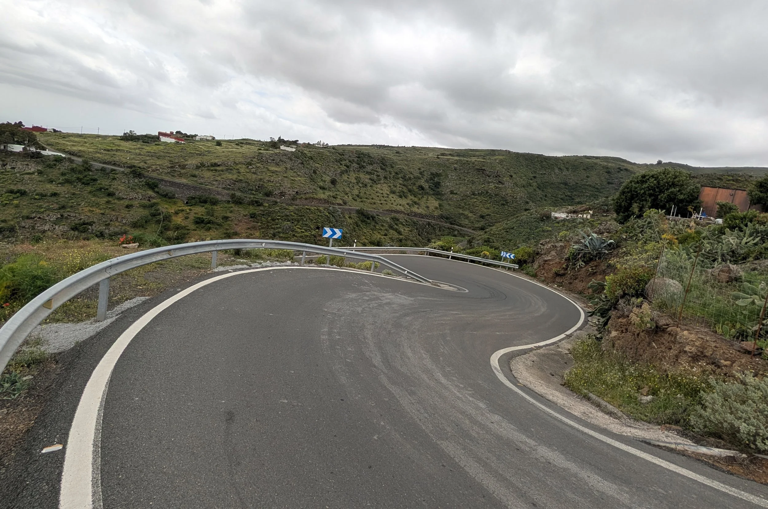 A winding mountain road with a metal guardrail, surrounded by green hills and shrubs under a cloudy sky.