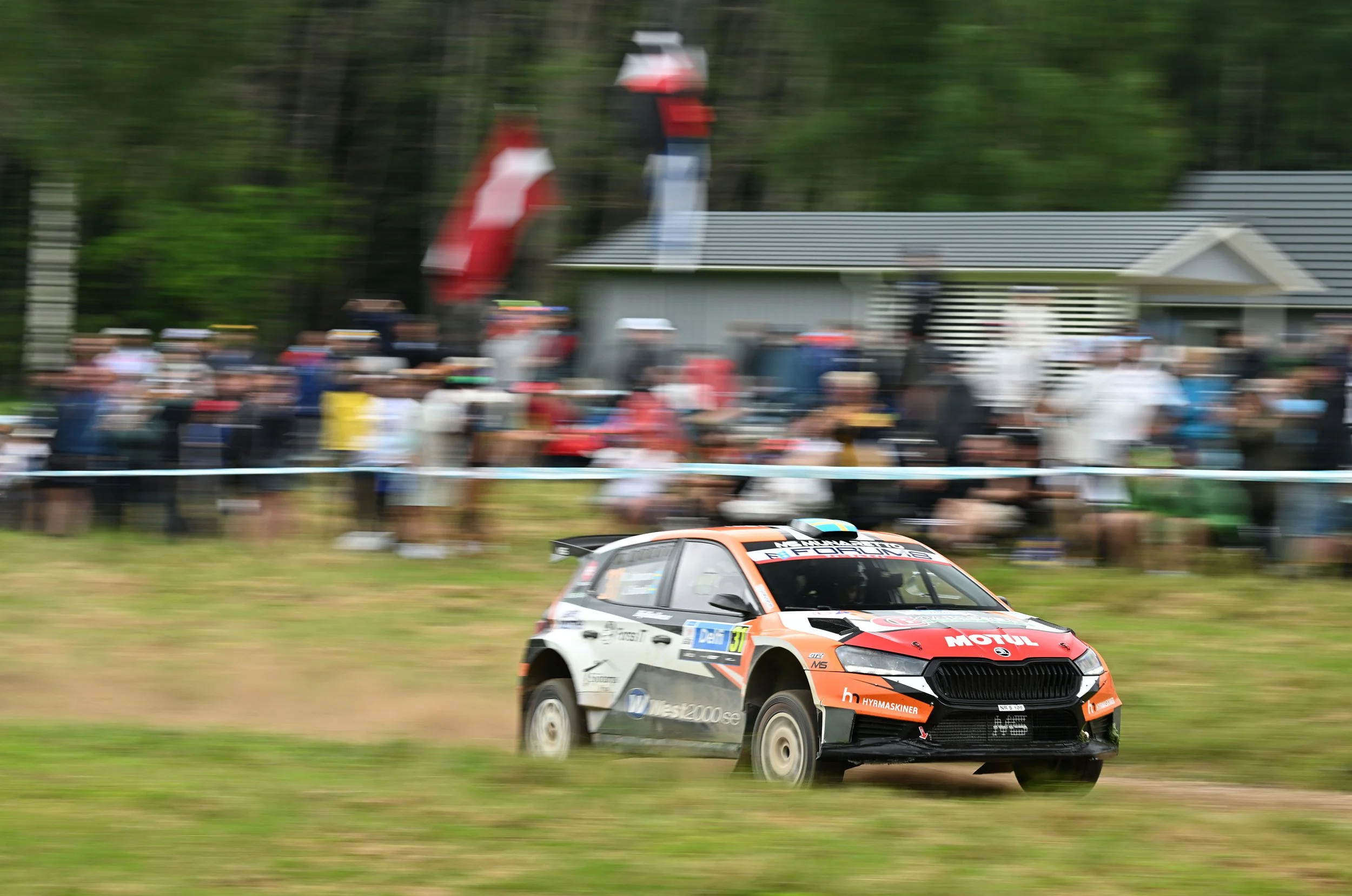 A rally car racing on a dirt track with a crowd of spectators watching from the side and a background of houses and trees.