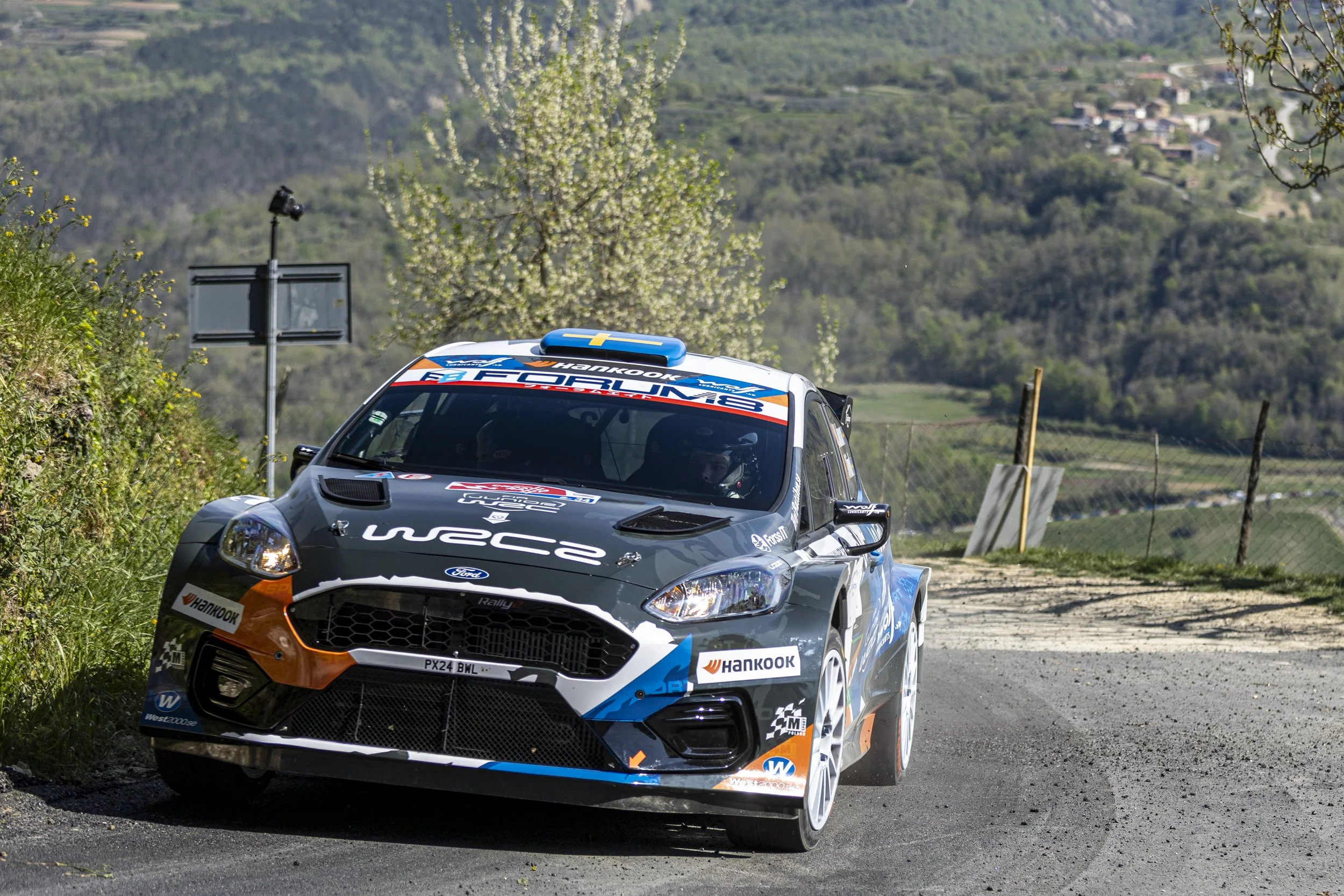 A Ford rally car on a dirt road with a rural hillside in the background, surrounded by flowering trees and a wire fence.