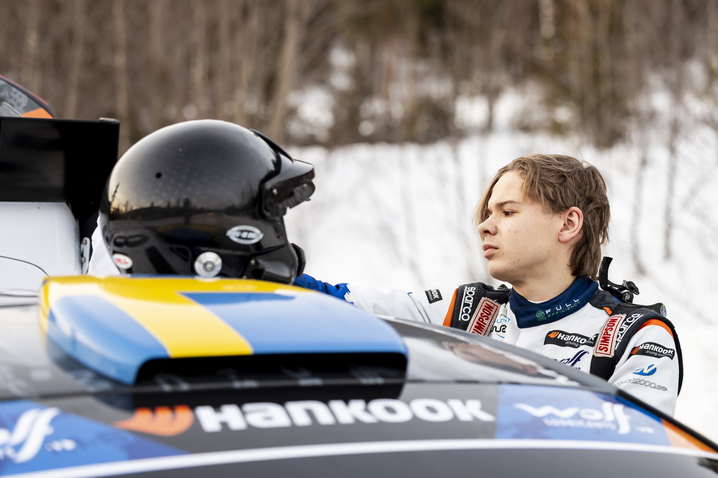 Young male race car driver in racing suit and gear looking at a racing helmet on a race car in a snowy outdoor setting with trees in the background.