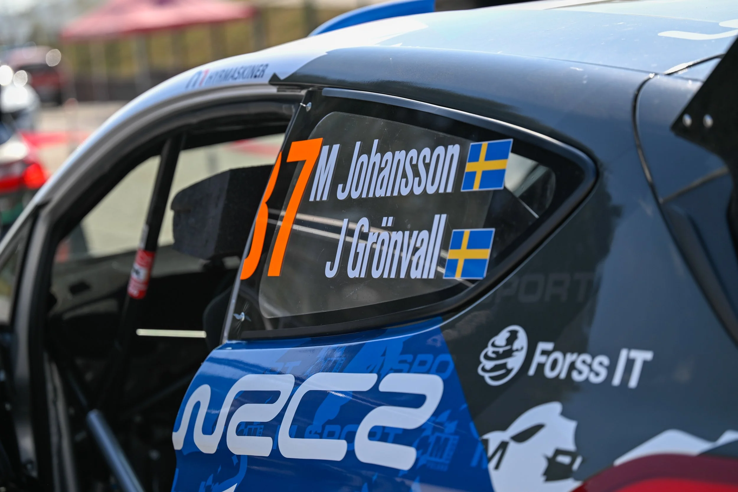 Close-up of a rally car's side window displaying the driver and co-driver's names, Johansson and Grönvall, along with their Swedish flags, and various sponsor stickers.