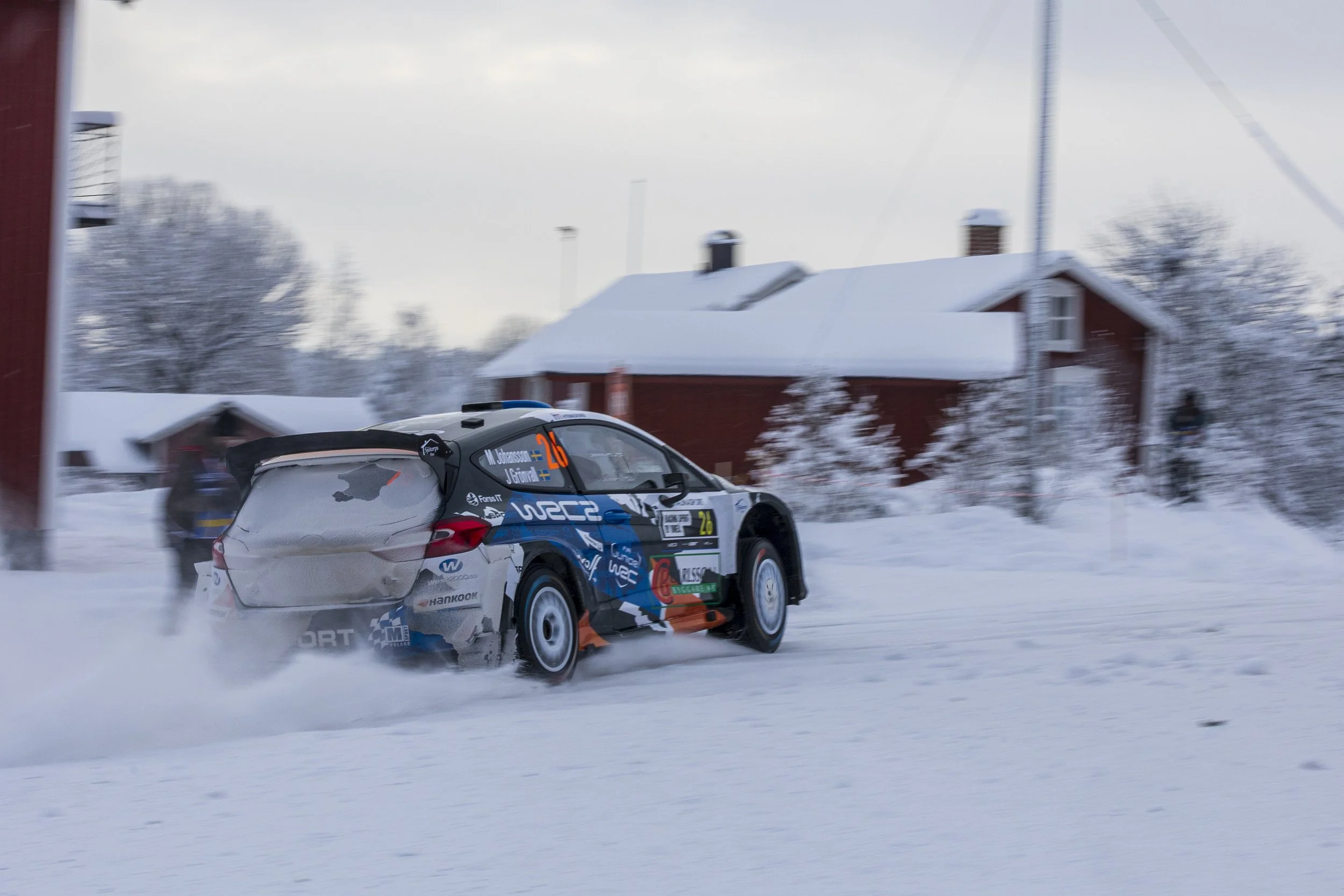 A rally car speeding on a snowy road with snow-covered houses and trees in the background.