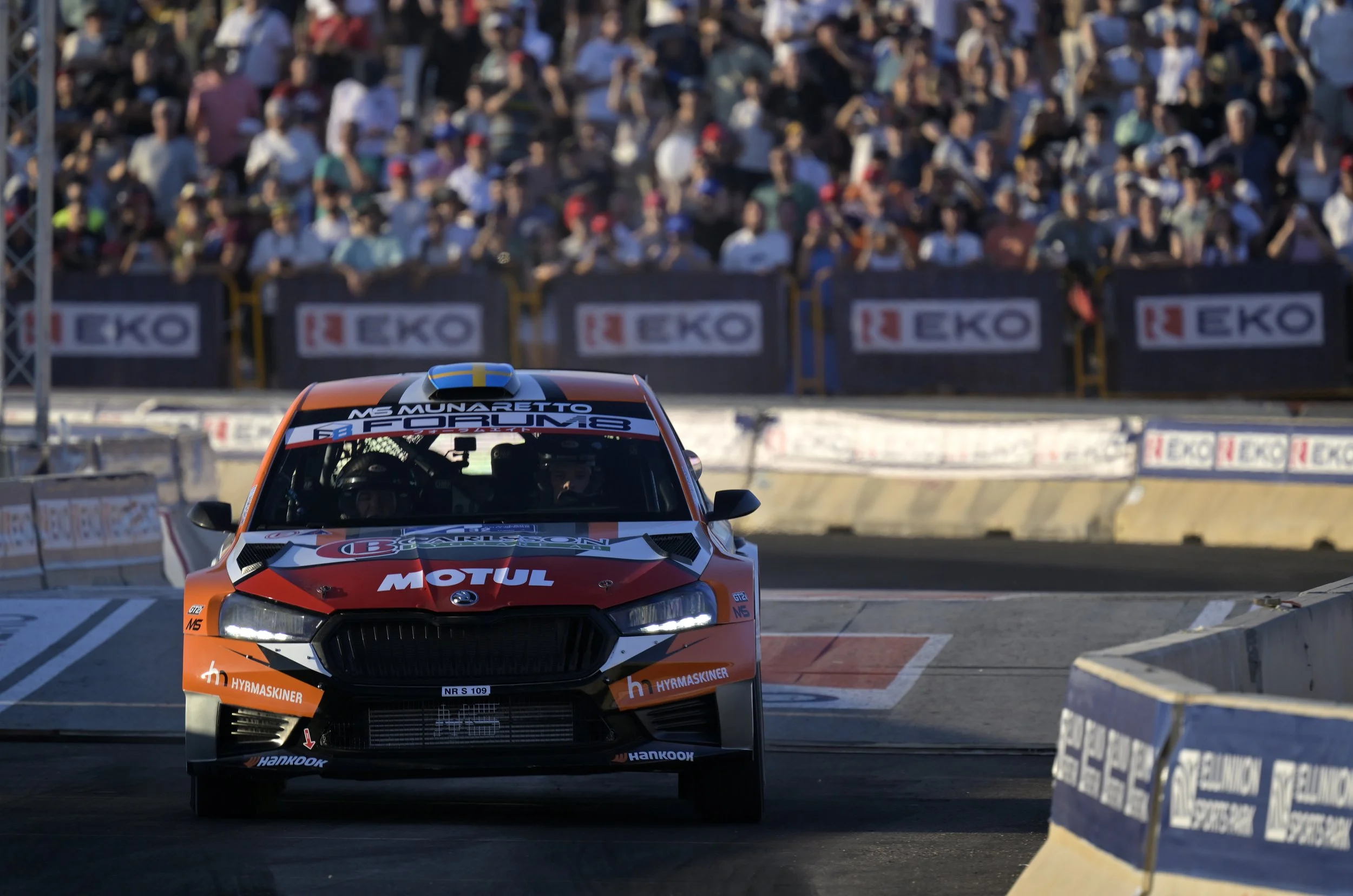 Orange and black rally car racing on a track with a crowd of spectators in the background.