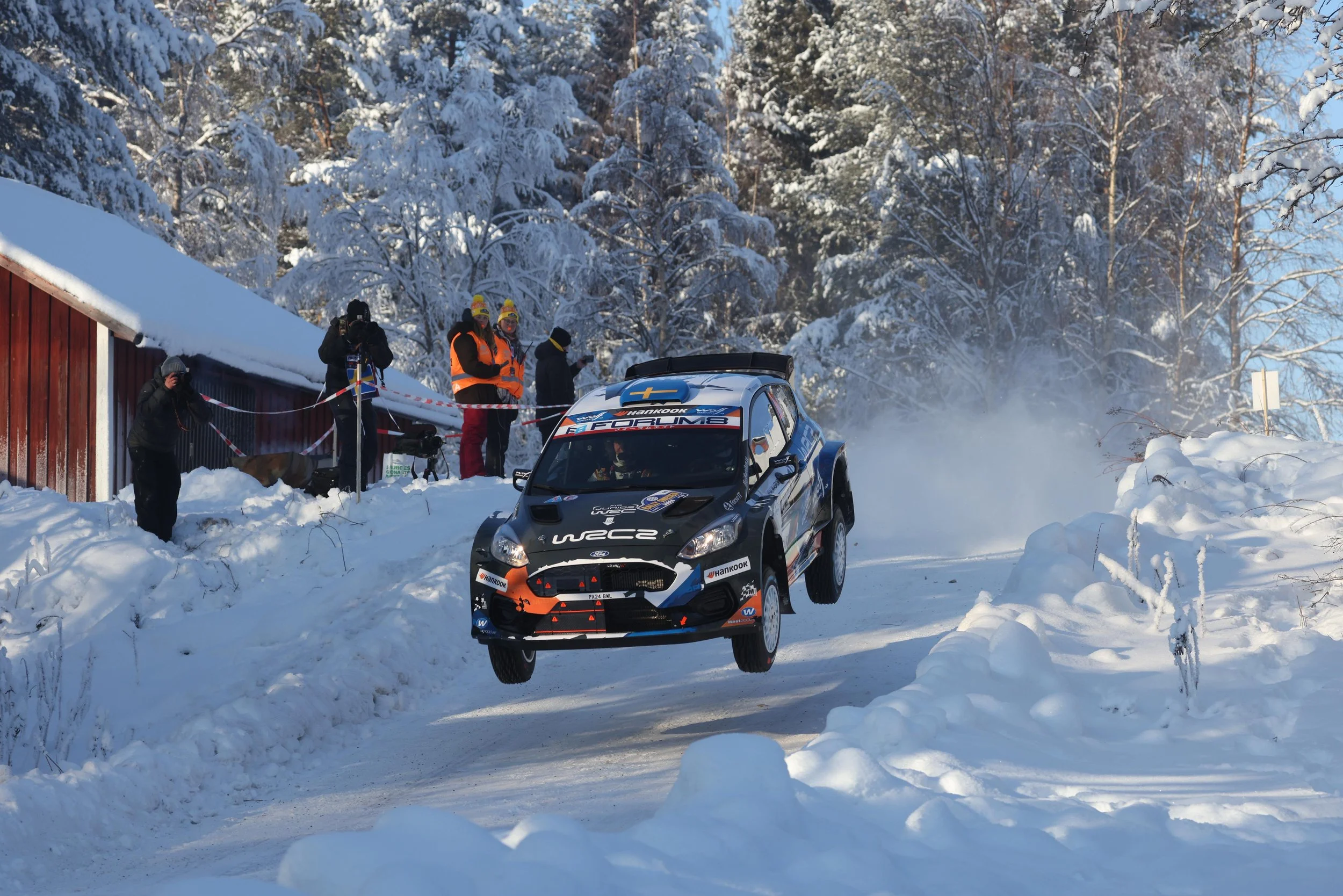 A blue and black rally car racing through snowy terrain with snow-covered trees in the background, and several people in winter gear watching nearby.