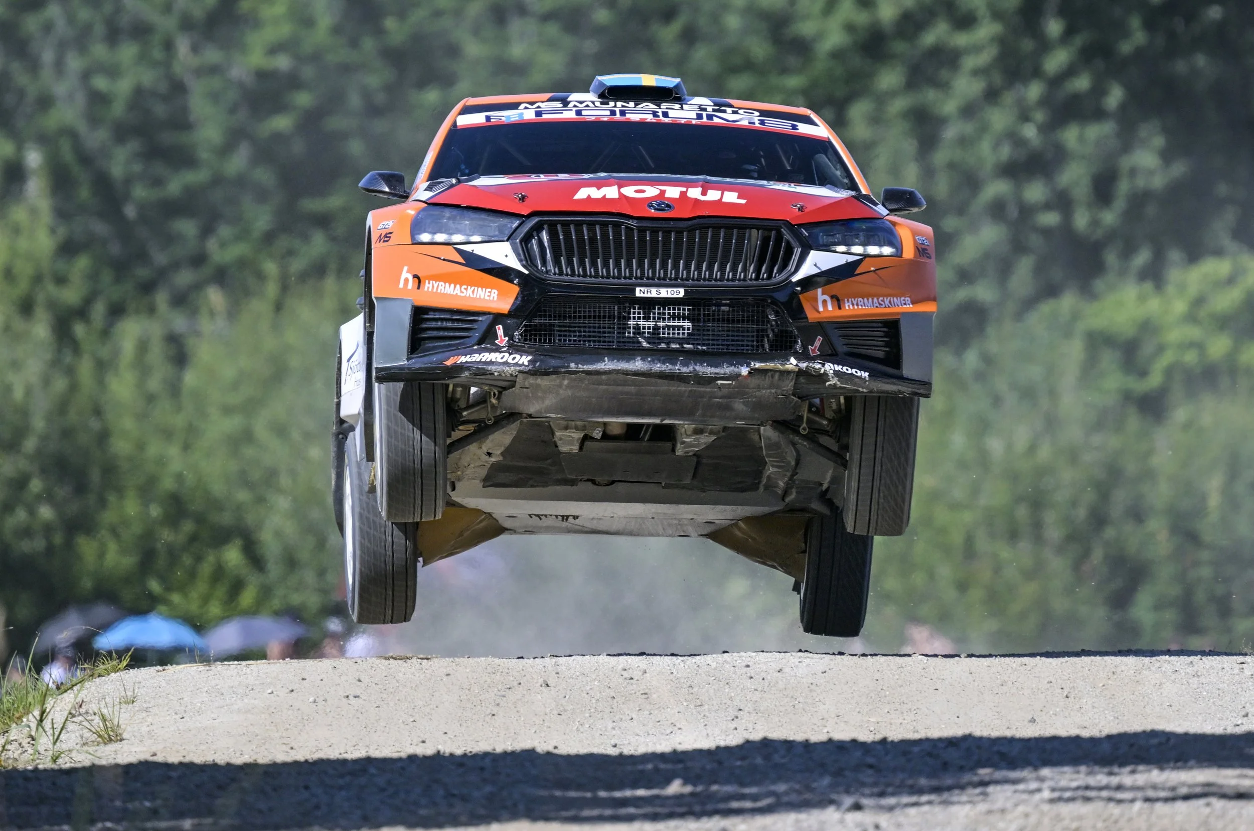 A racing car in mid-air during a race, with a green wooded background, racing decals, and sponsorship stickers on the car.