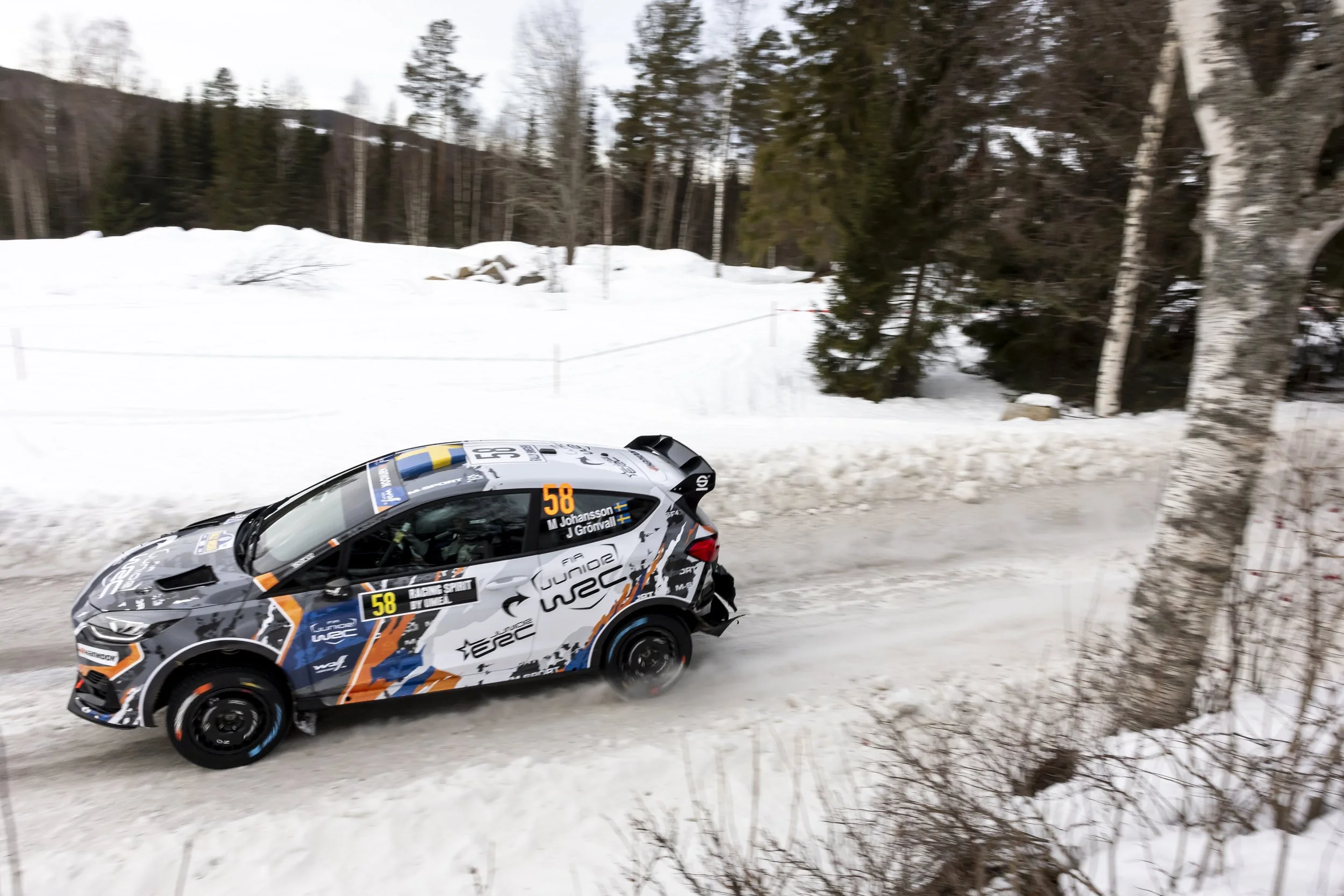 A rally car racing on a snowy road in a winter landscape with tall trees and rolling snow-covered terrain.