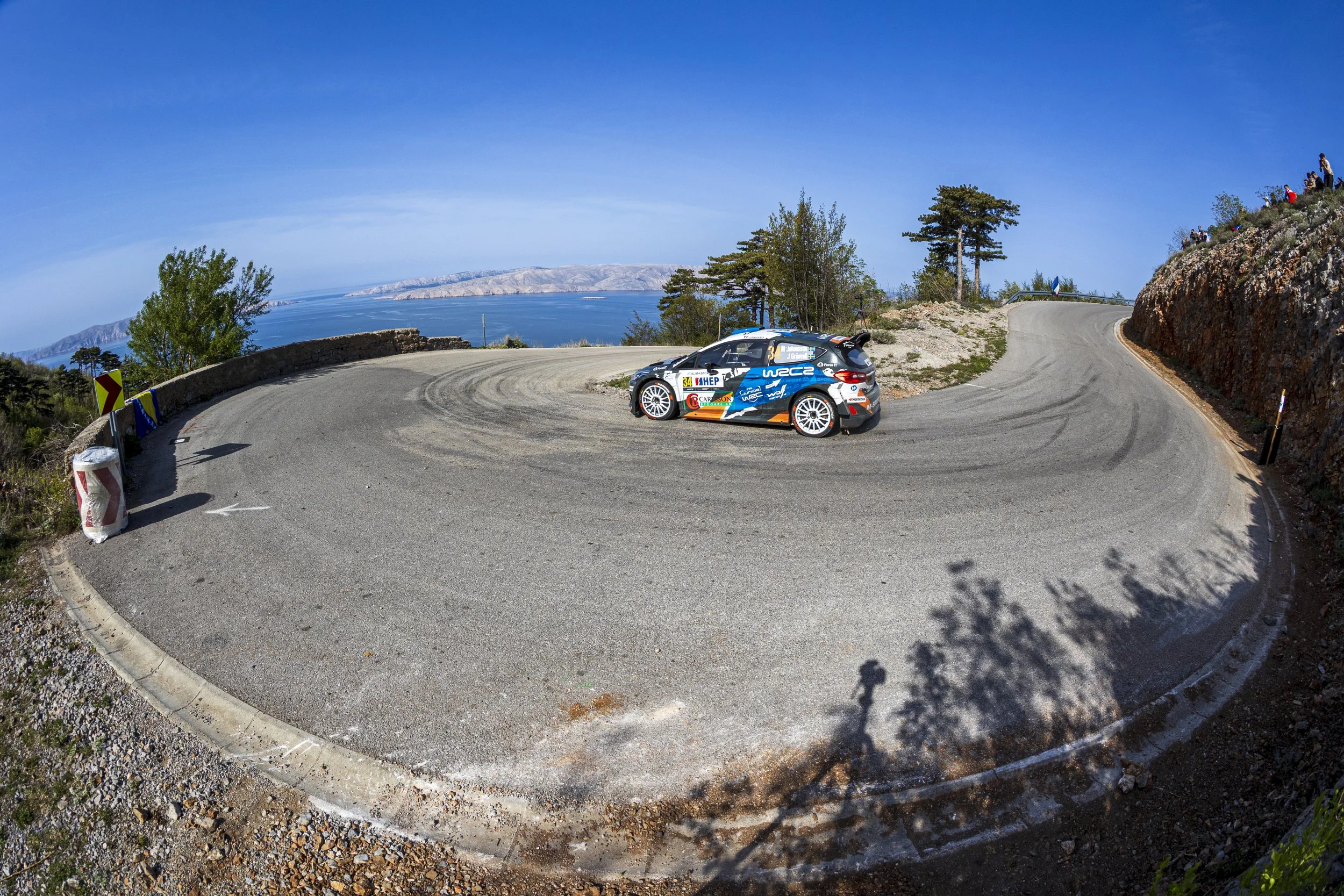 A rally car driving around a sharp curve on a mountain road with a scenic view of water and land in the background.
