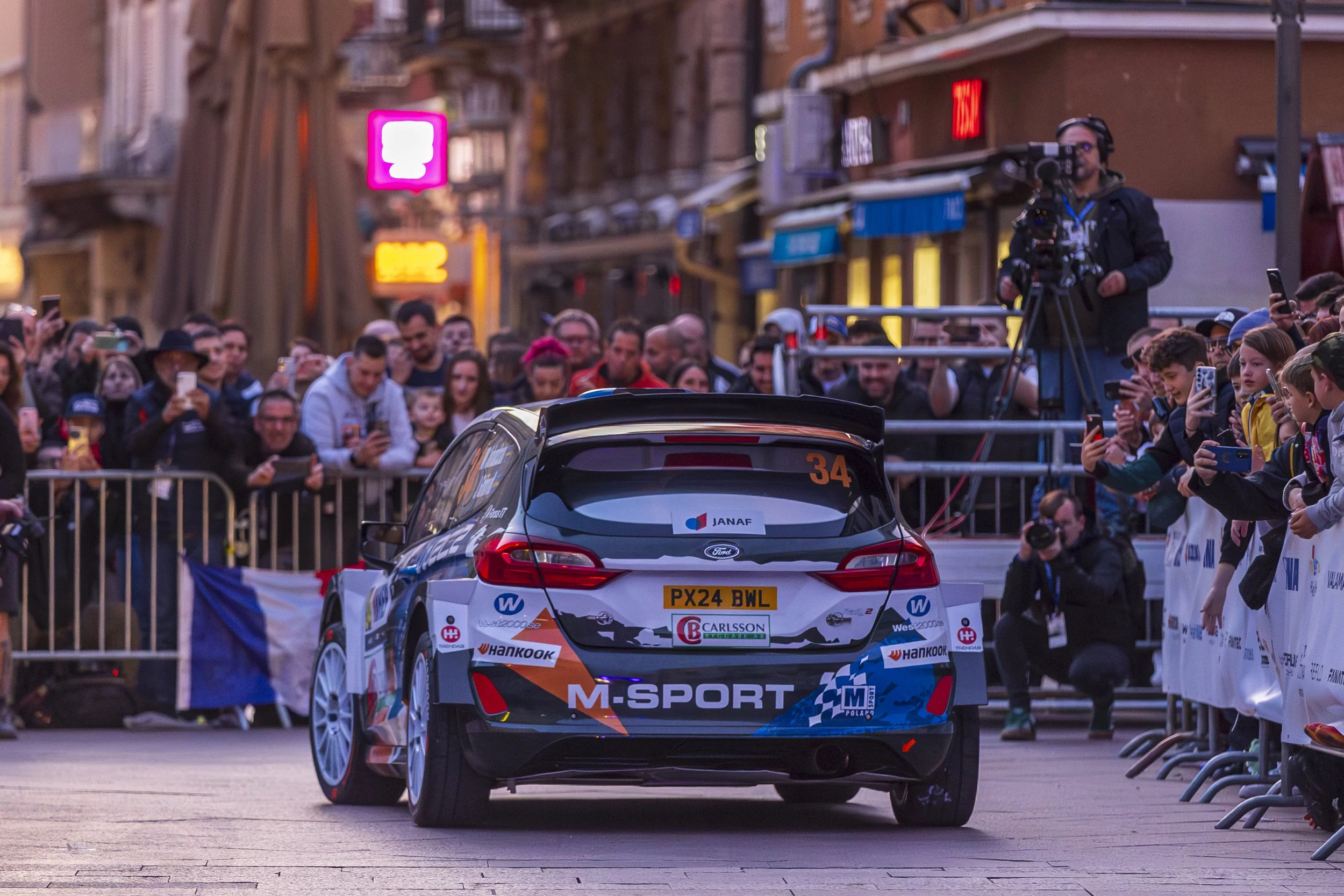 A rally car on a city street lined with spectators behind barriers during a race event.