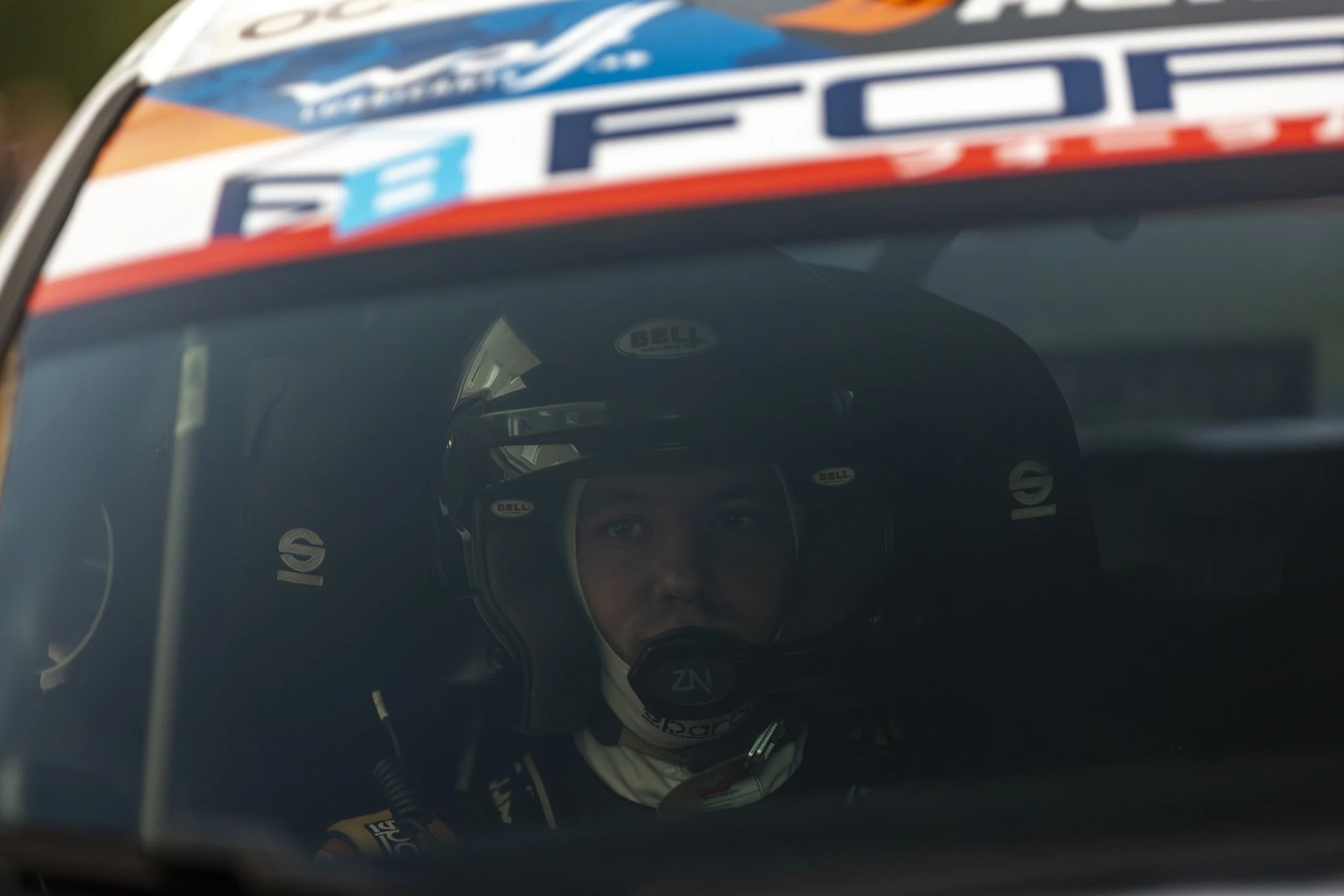 Close-up of a race car driver wearing a racing helmet and suit, sitting inside a race car, with the driver looking through the windshield.