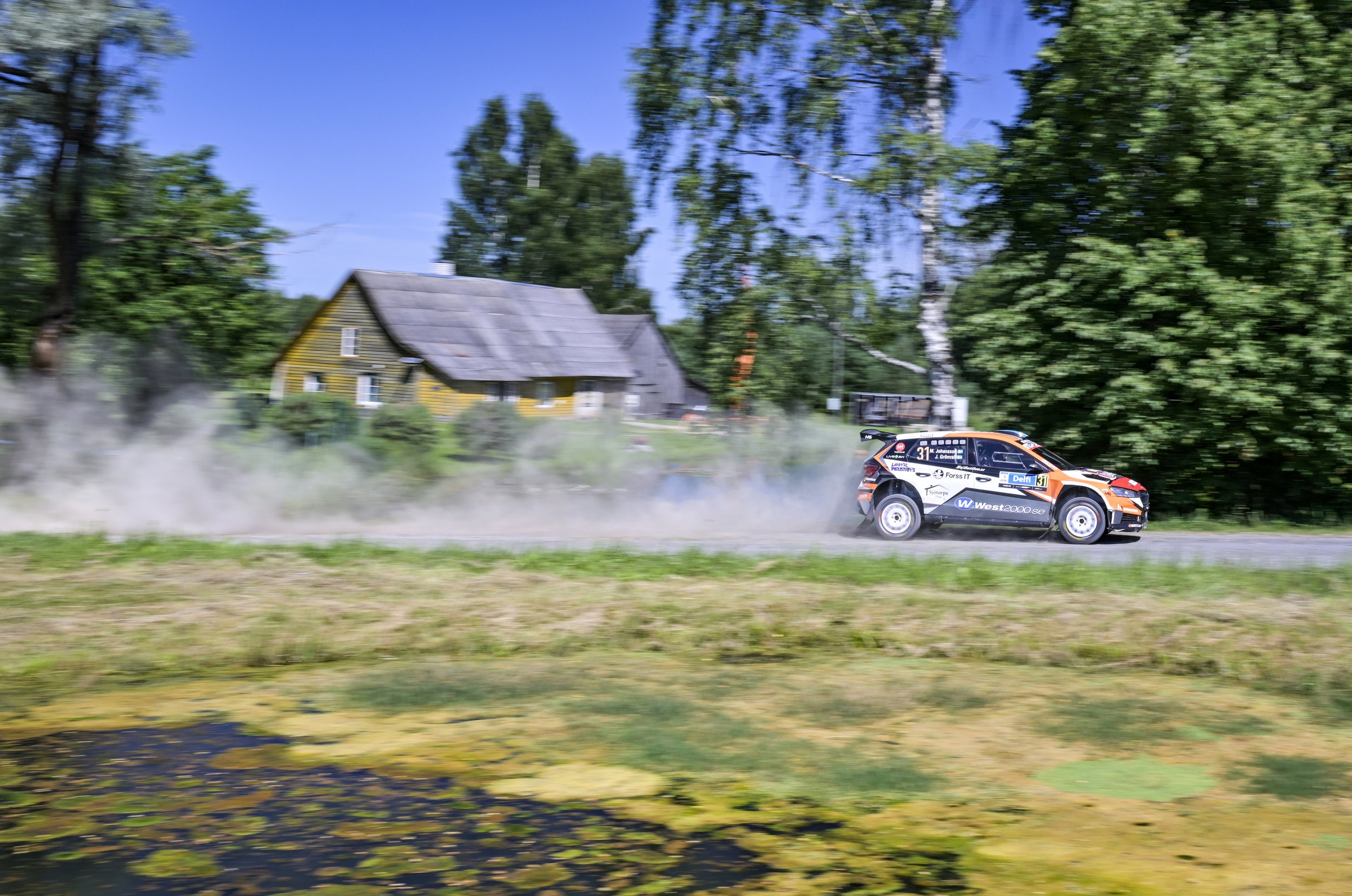 A rally car speeding along a rural dirt road, kicking up dust, with green trees and a house in the background under a clear blue sky.