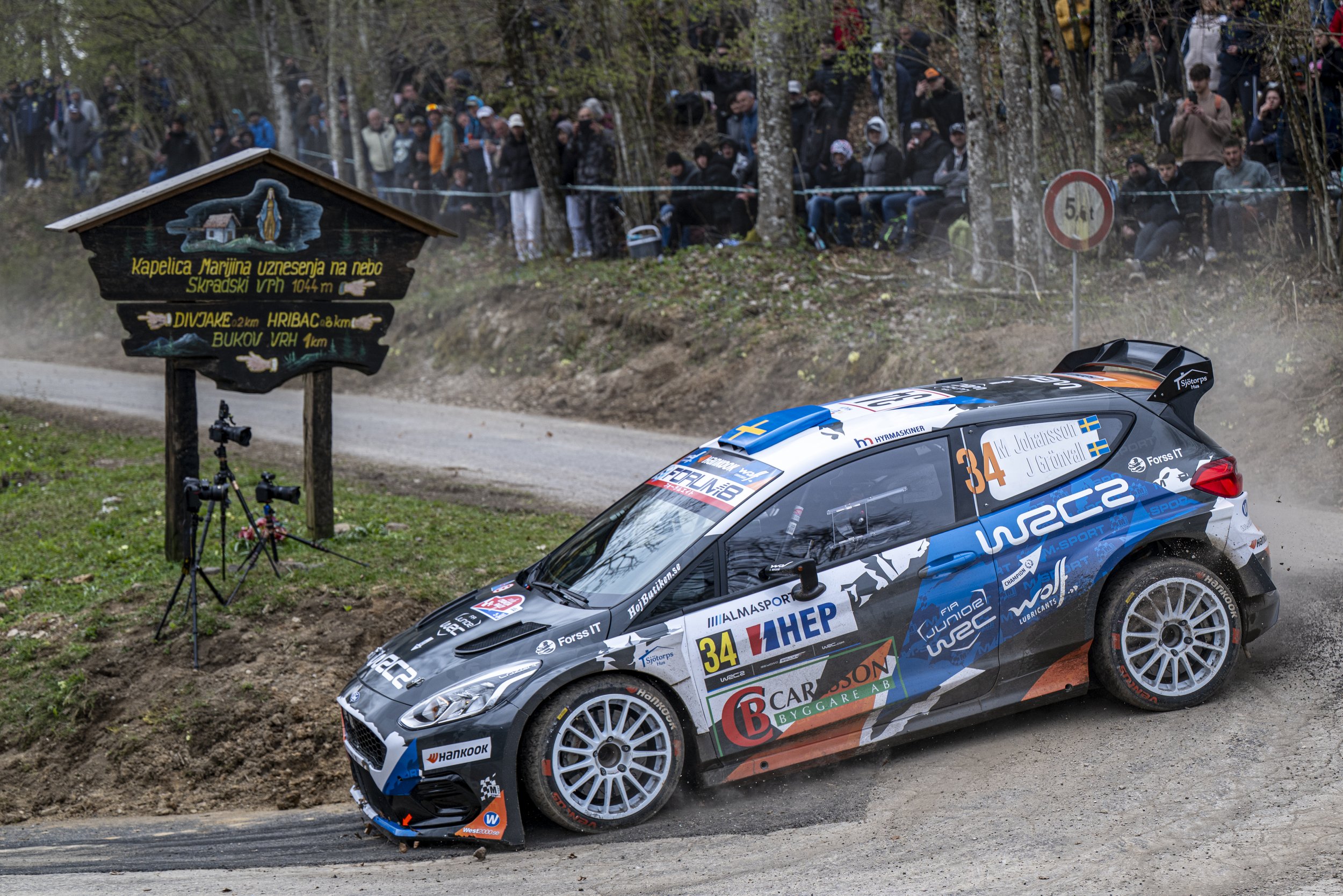 A rally car with sponsor logos racing on a dirt road, kicking up dust, with a crowd of spectators behind a safety barrier and a signpost with directions in the background.