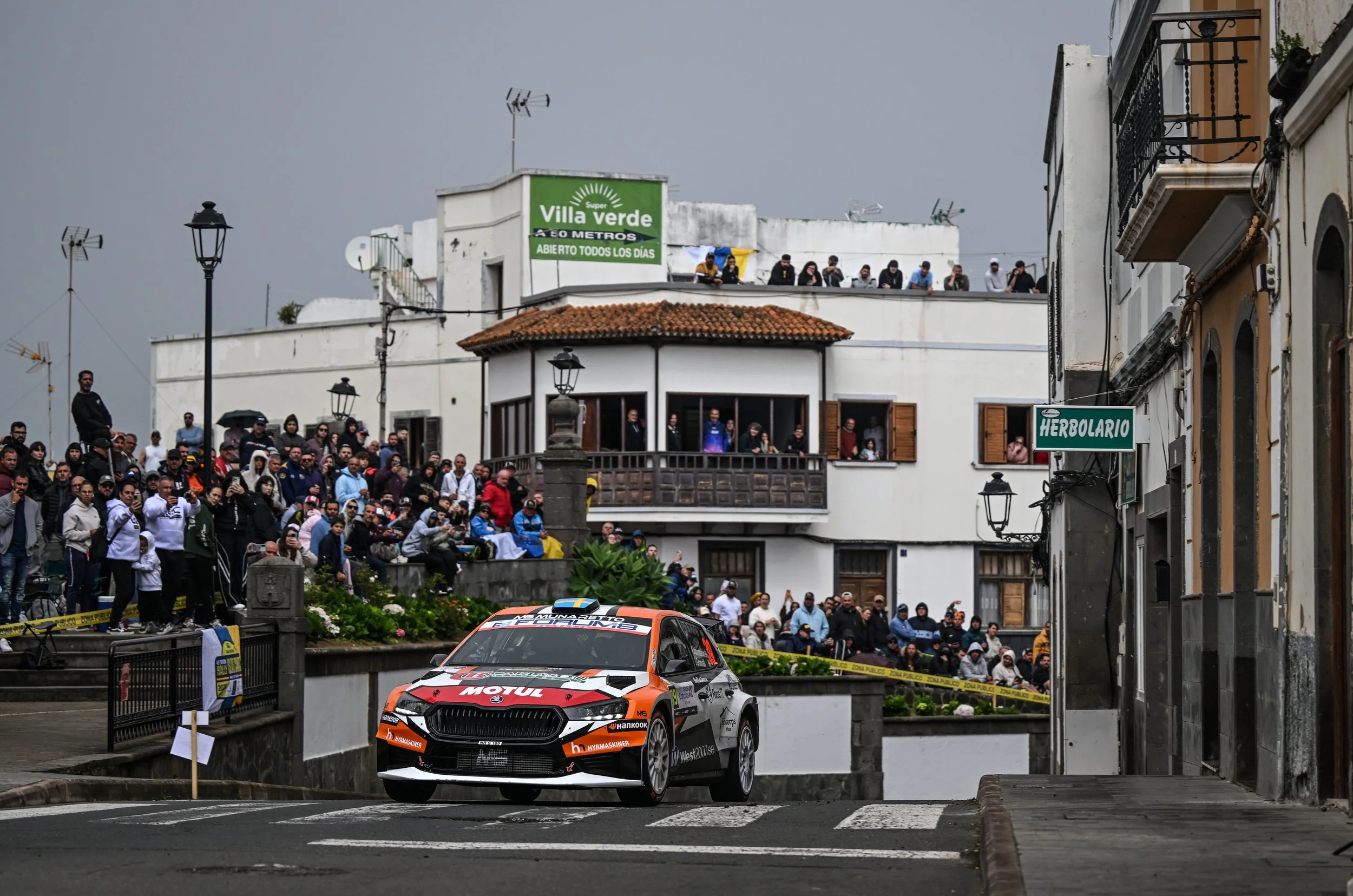 A rally car racing through a city street with spectators on the sidewalks and rooftops, with buildings, streetlights, and a cloudy sky in the background.