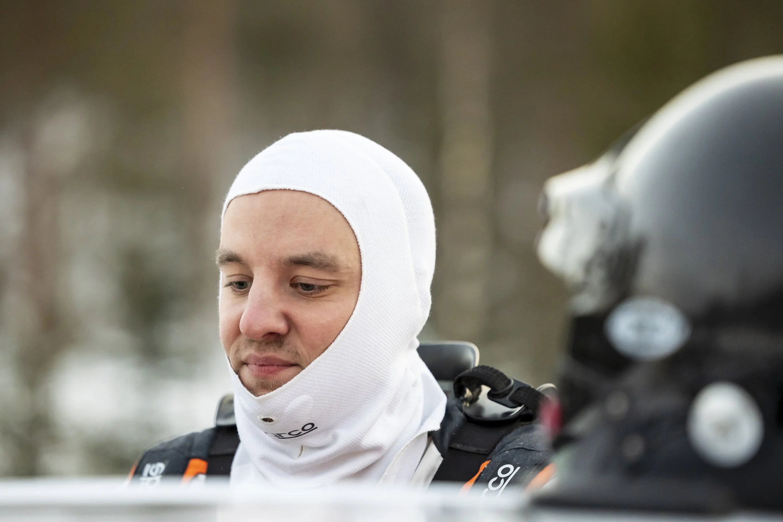 A man wearing a white hat and a neck gaiter, dressed in outdoor gear, is focused on an activity outdoors, with trees blurred in the background.