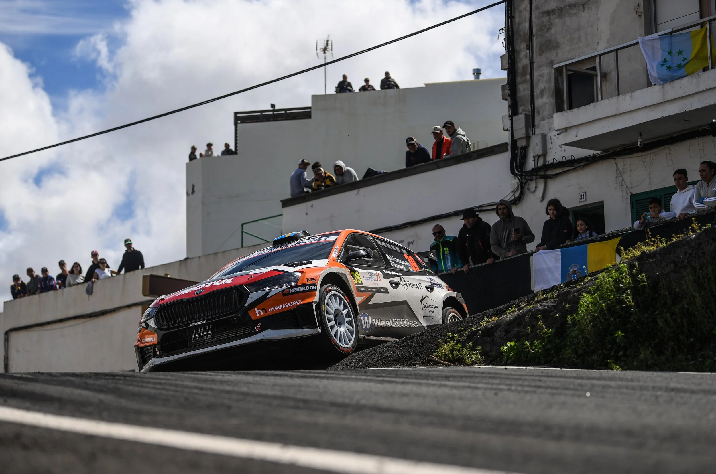 A rally car racing on a steep downhill road with spectators watching from a balcony and street level.