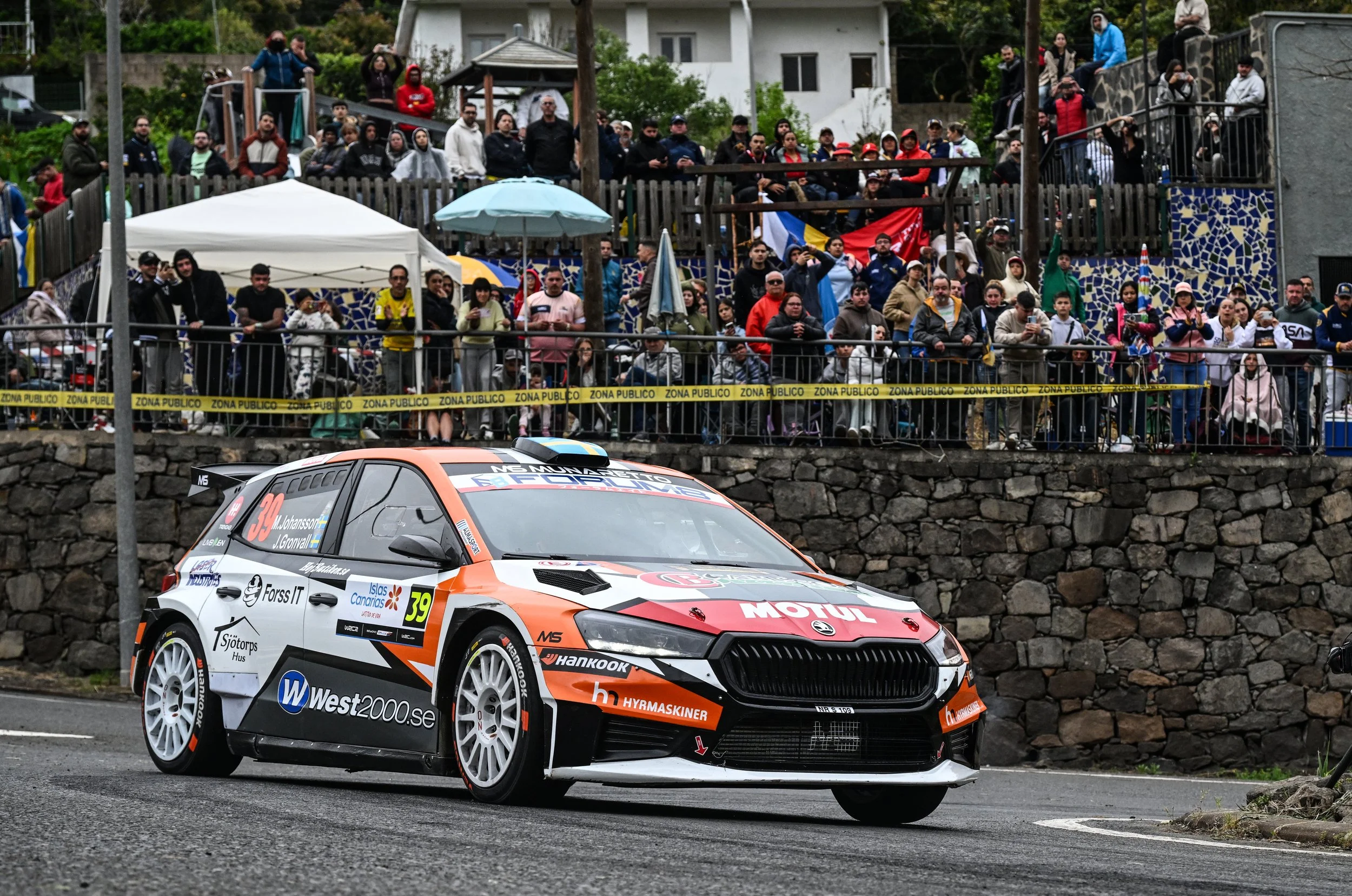 A rally car on a road with spectators behind a yellow 'ZONA PUBLICO' tape, viewers standing on a second-story balcony and ground level, some taking photos and wearing jackets, over a stone wall with houses and trees in the background.