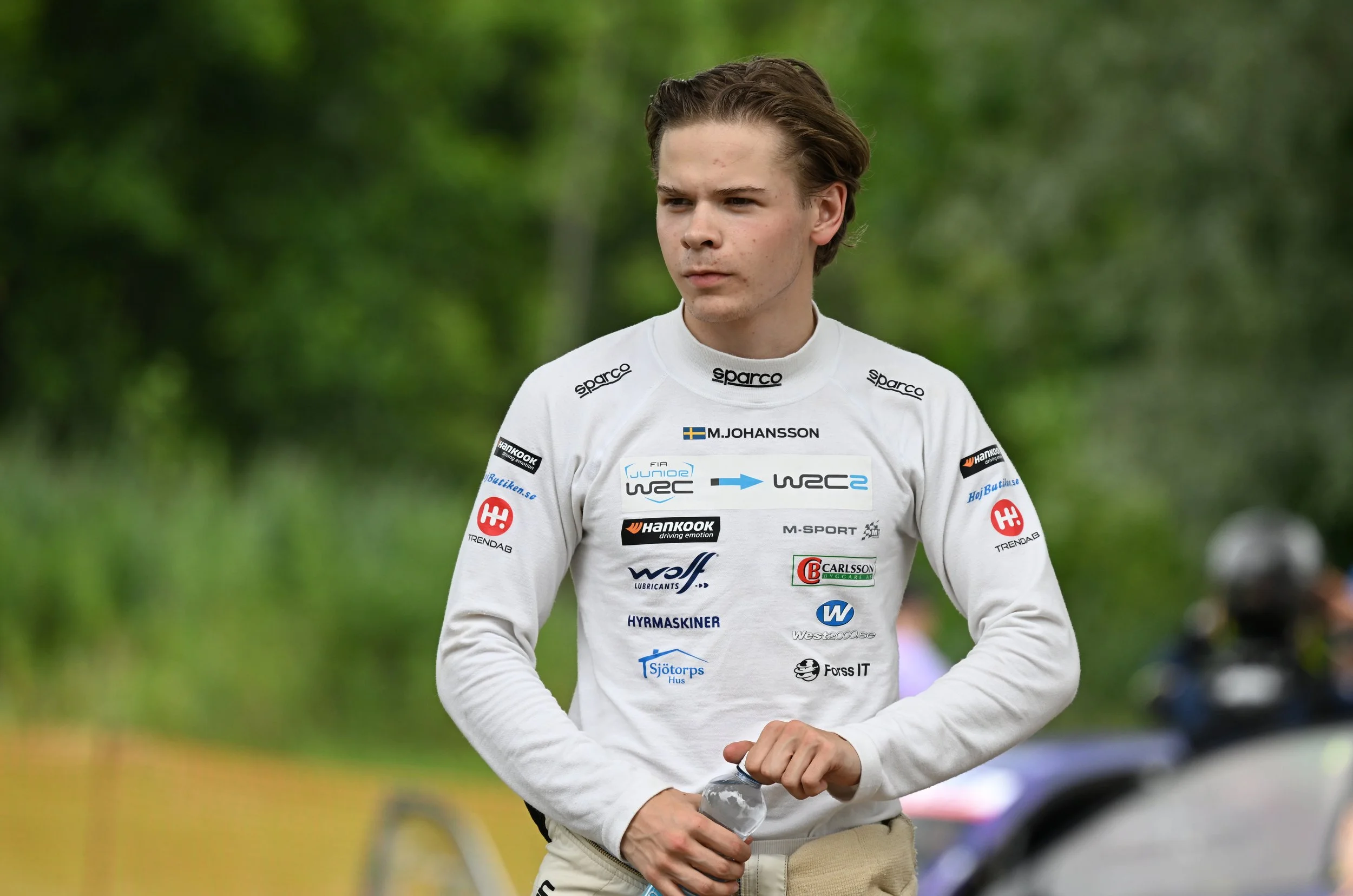 Young man with light skin and brown hair, wearing a white racing shirt with various sponsor logos and the name M. Johansson, holding a plastic water bottle, standing outdoors in a green, wooded area.