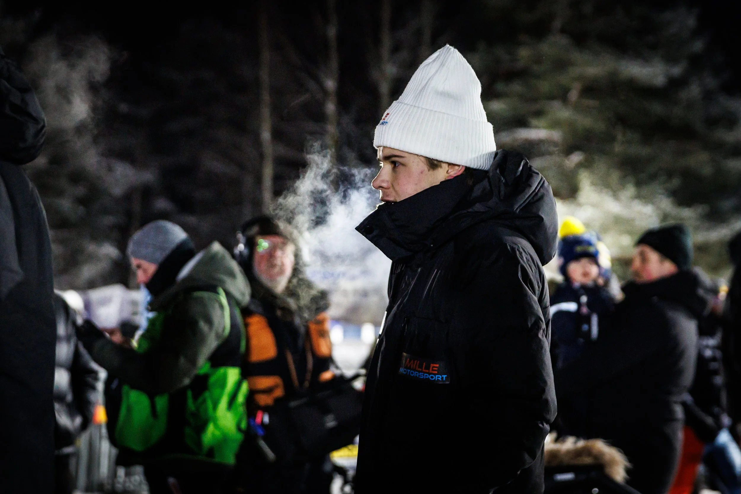 Young man wearing a white beanie and black winter jacket at an outdoor nighttime gathering with several other people in winter clothing.