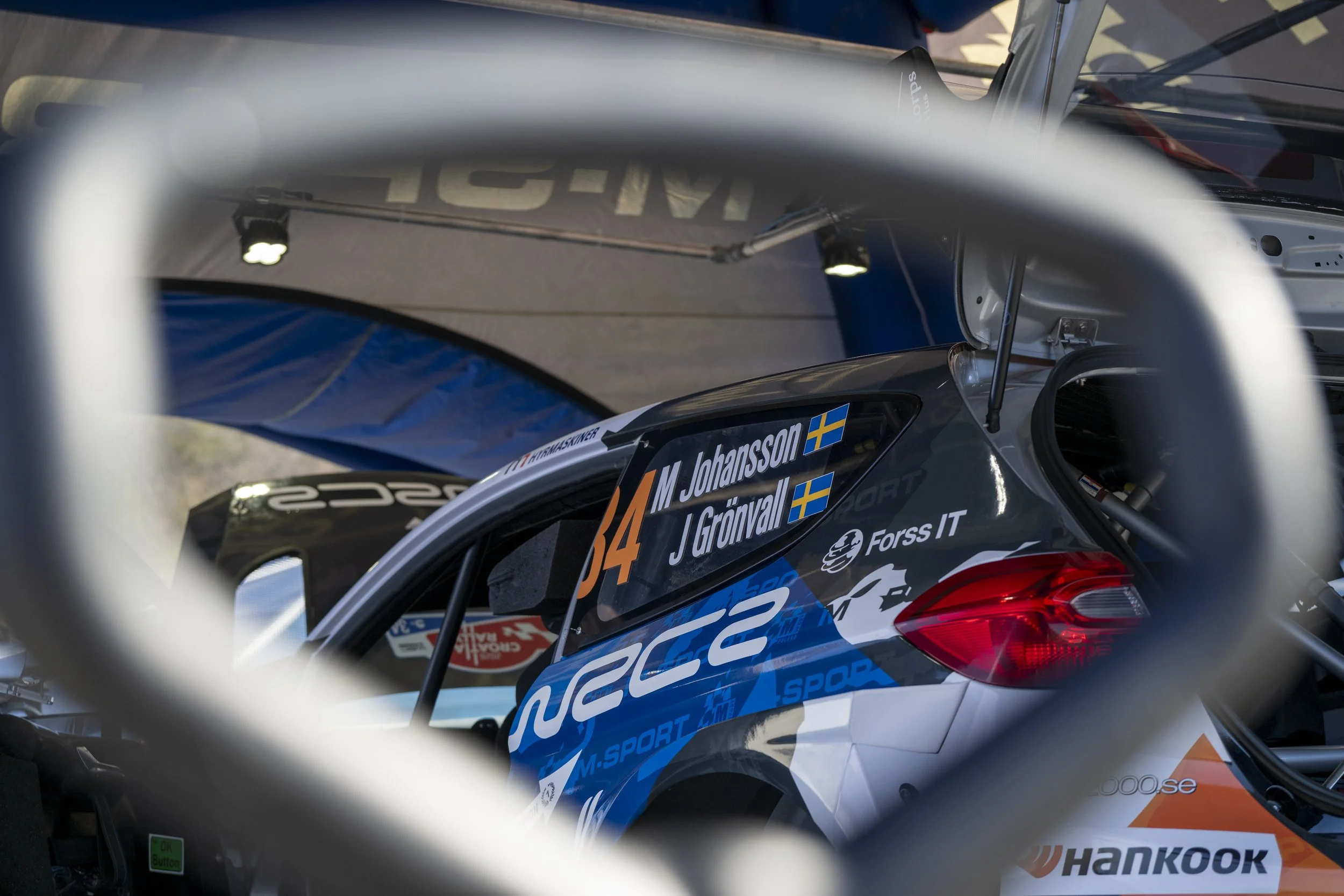 Close-up of a race car through a white metal barrier, showing the car's windshield, side window, and rear wing with racing decals and the Swedish flag.