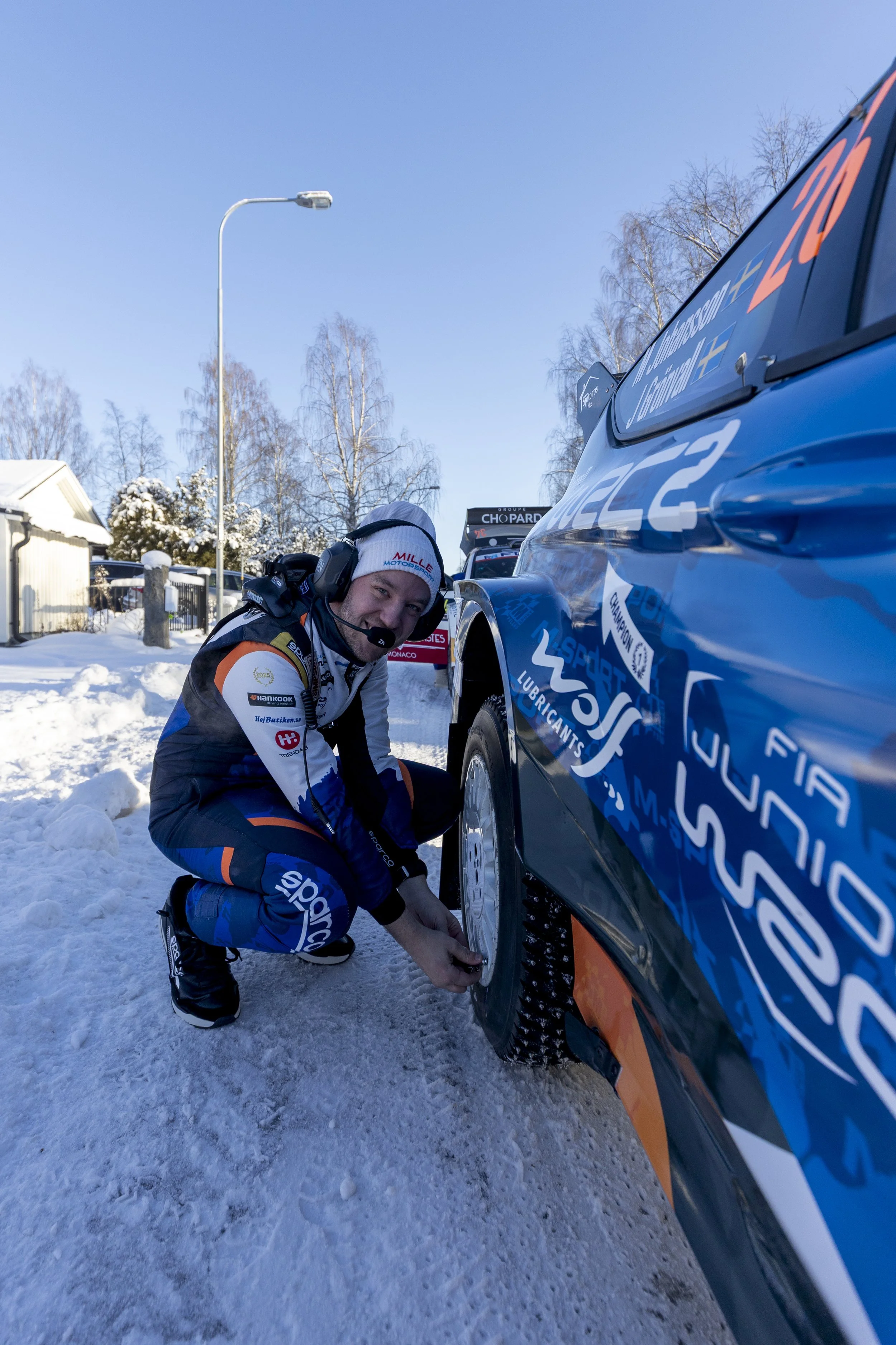 A male race car driver in racing gear crouches by a blue race car on a snowy outdoor track, working on the tire.