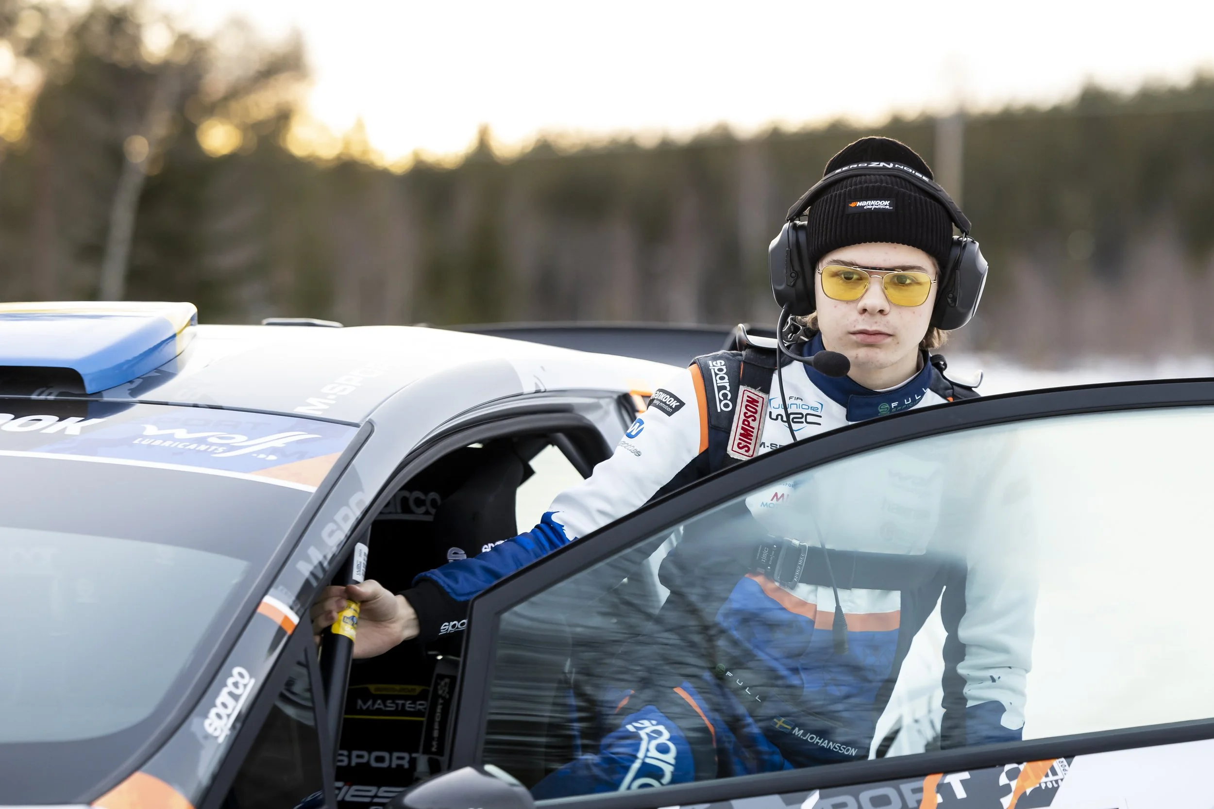 A young male race car driver wearing a black beanie, yellow-tinted glasses, and a racing suit with various sponsor patches, standing next to his cockpit during daytime in an outdoor setting with blurred trees in the background.