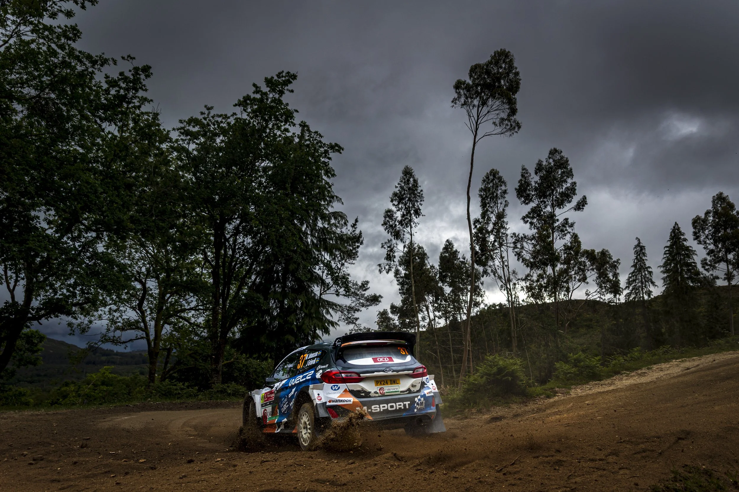 A rally car racing on a dirt track under dark, cloudy skies, surrounded by tall trees and hilly terrain.