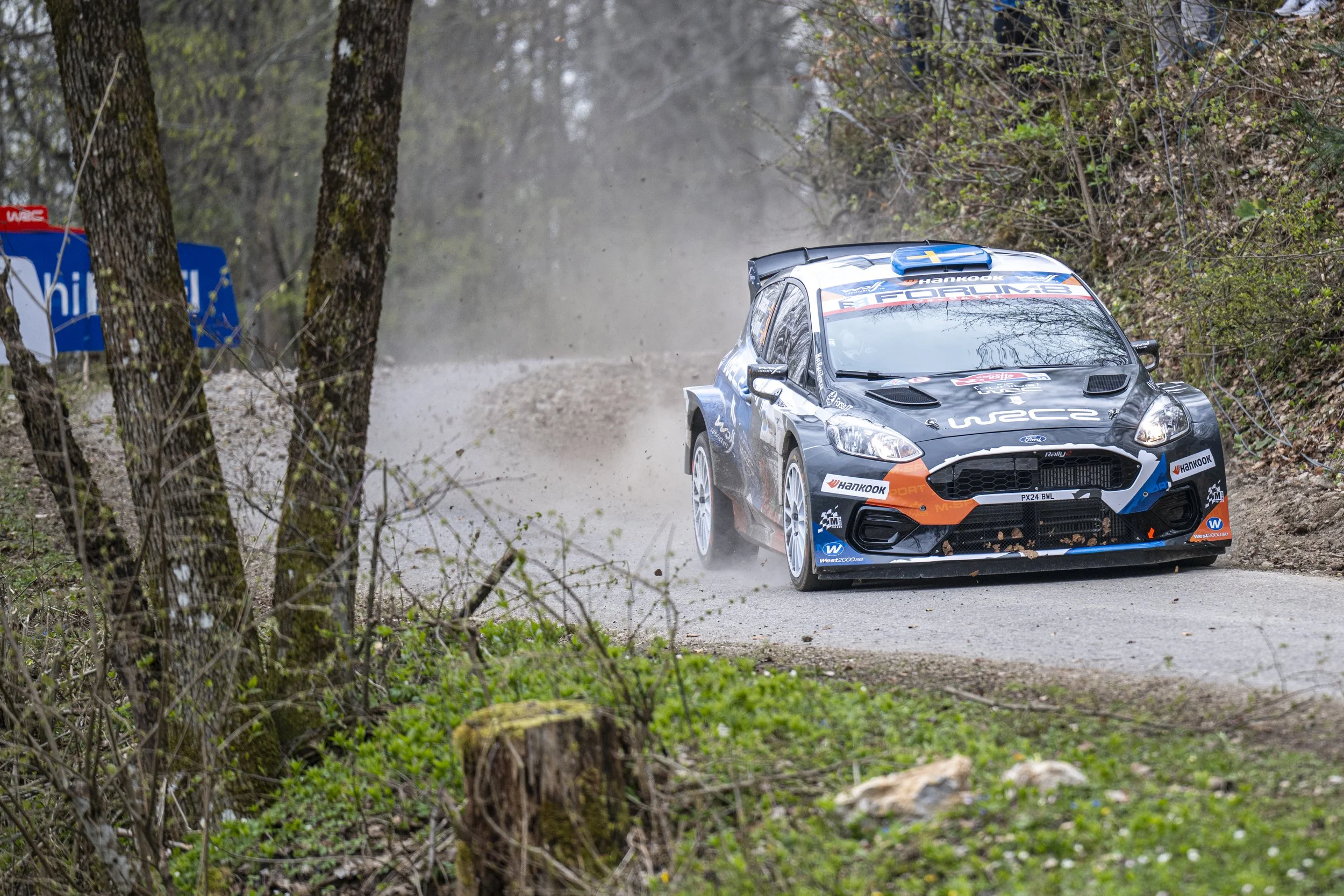 A rally car speeding on a forested dirt and paved road, kicking up dust, with trees and foliage on either side.
