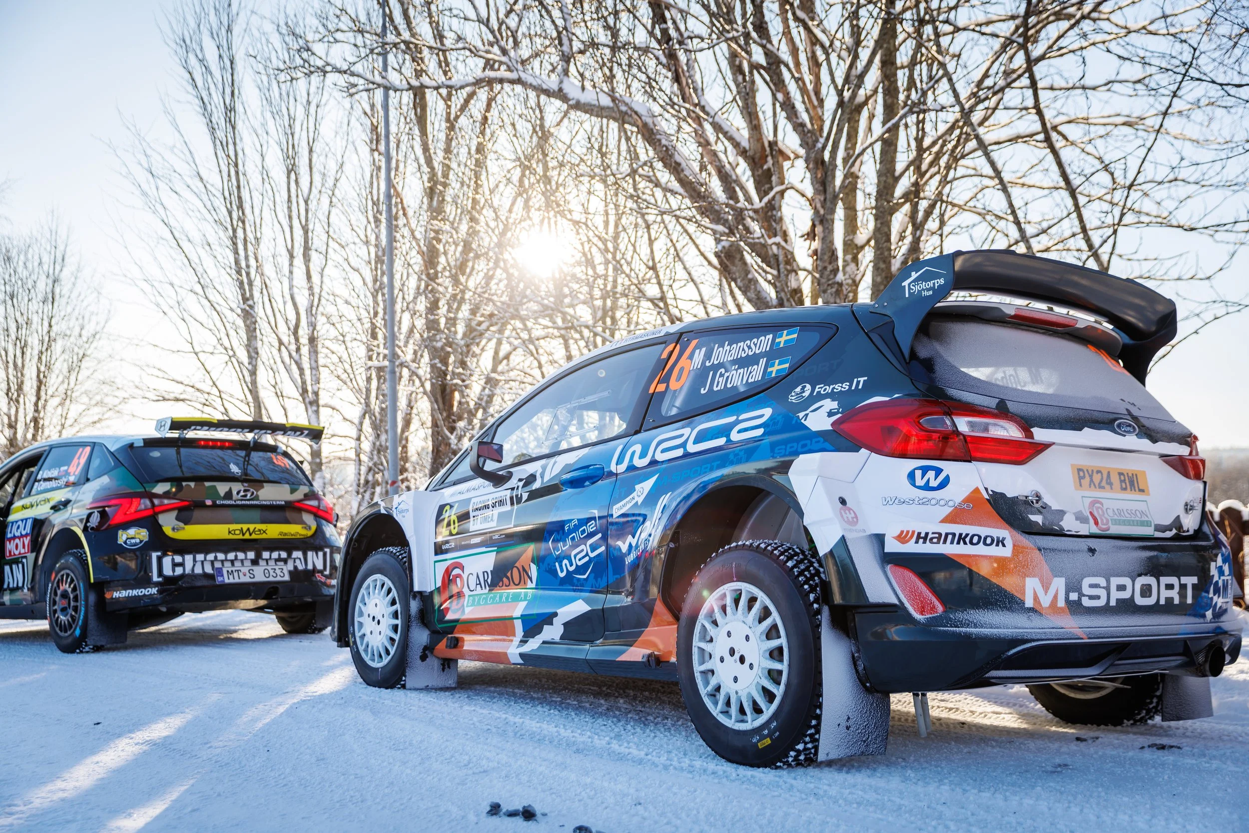 Snow-covered rally cars with racing sponsorship logos parked outdoors against a background of trees and a bright sky.
