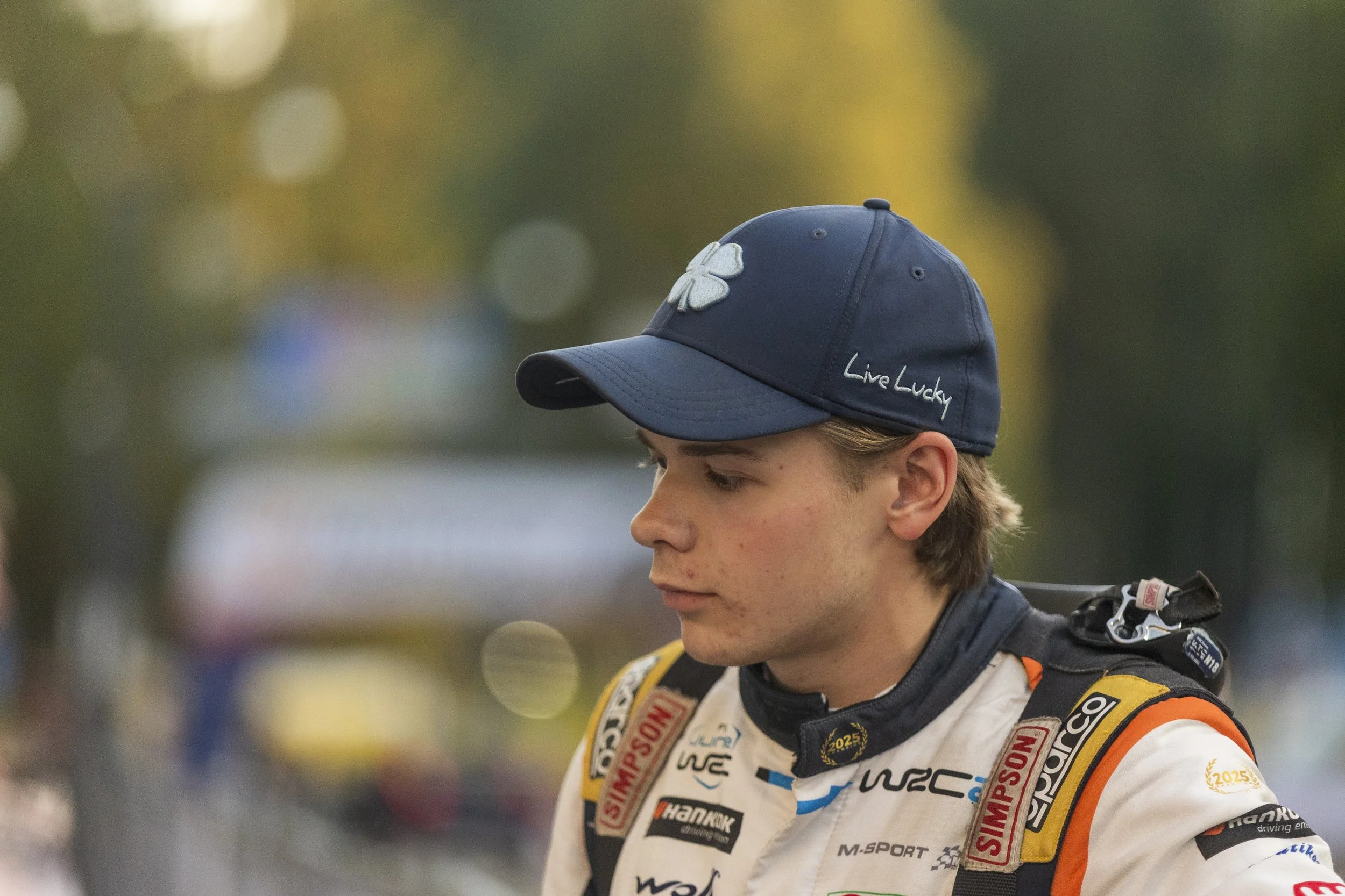 A person wearing a dark blue baseball cap with a white four-leaf clover and the words "Live Lucky" embroidered on it. The individual is dressed in a racing suit with various sponsor patches and is likely a race car driver. The background is blurred w