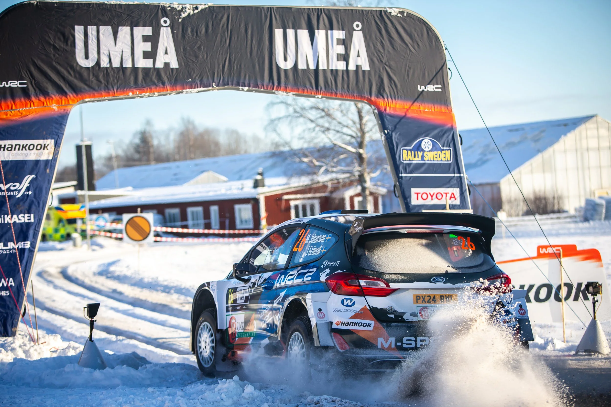 A rally car with the number 26 and driver's names J. Johnson and J. Granholm on the side, racing on a snowy track under a large UMEA banner during a winter rally event.