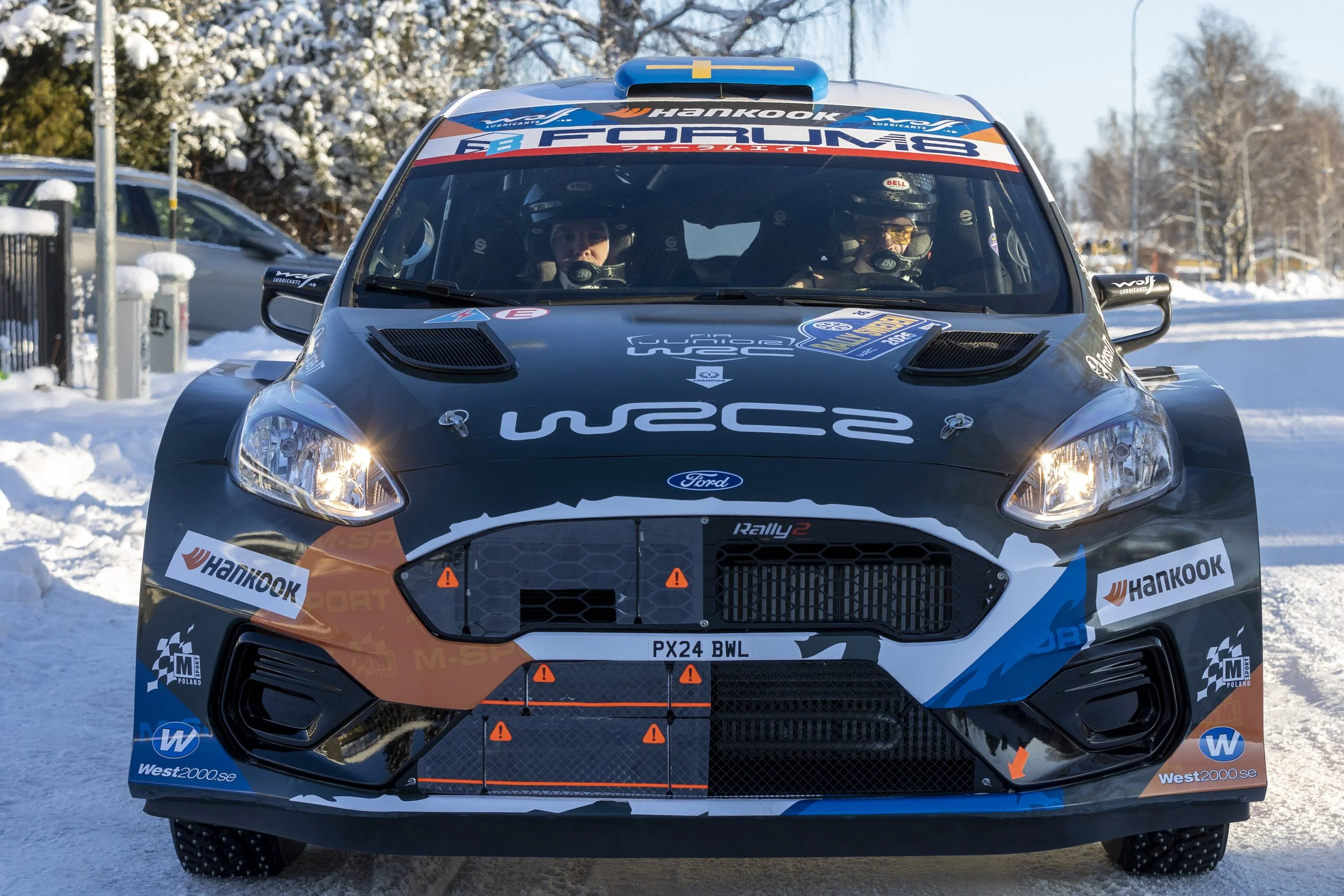 A rally car with blue, black, and orange livery is on snow, with two drivers inside wearing helmets and racing gear. The car has branding including Hankook, Ford, and WRC2, and a logo for West2000.se. Snow-covered trees and parked cars are visible in