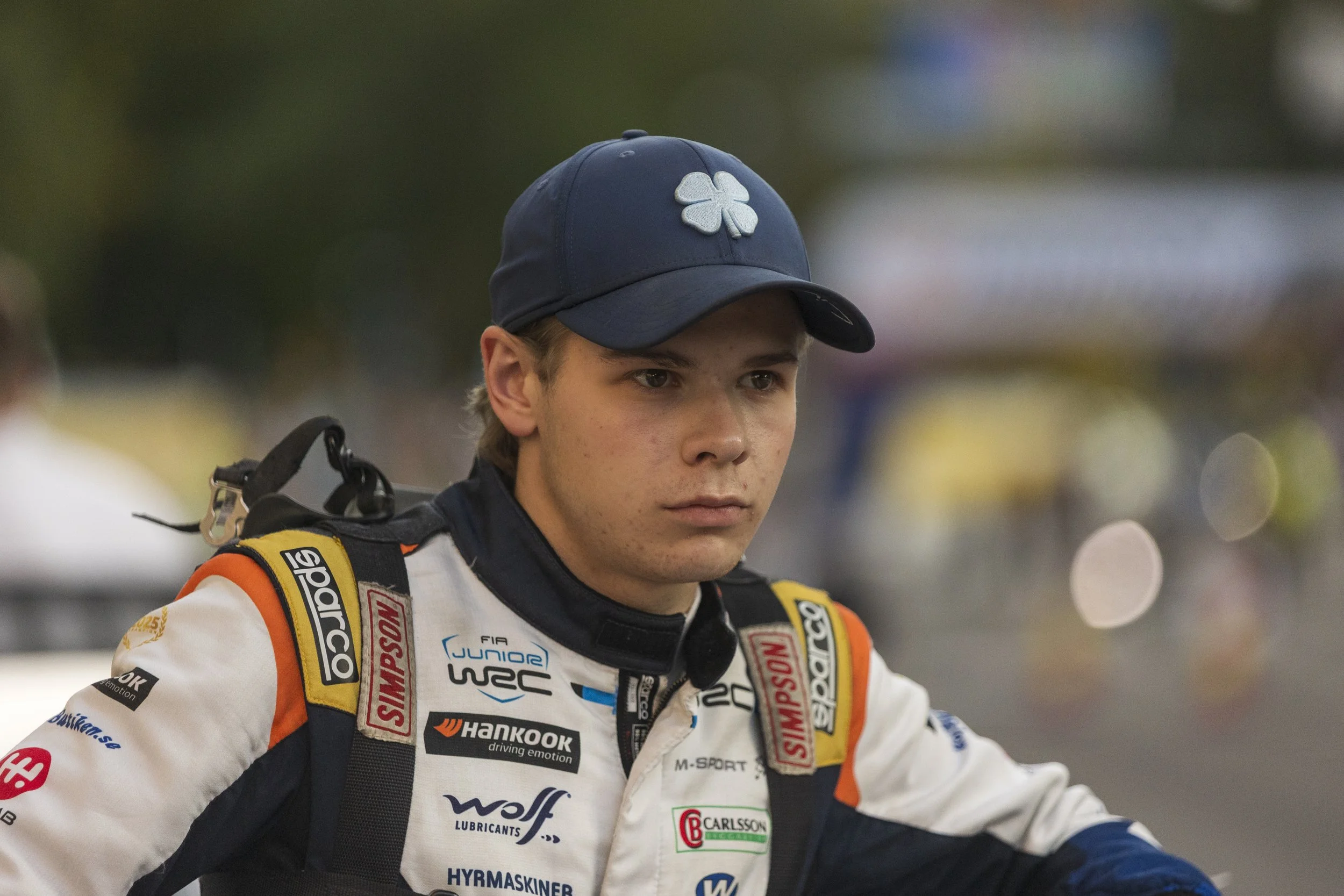 A young male race car driver in a racing suit and cap, looking serious, with various sponsor logos and patches on his suit, standing outdoors in a blurred background.
