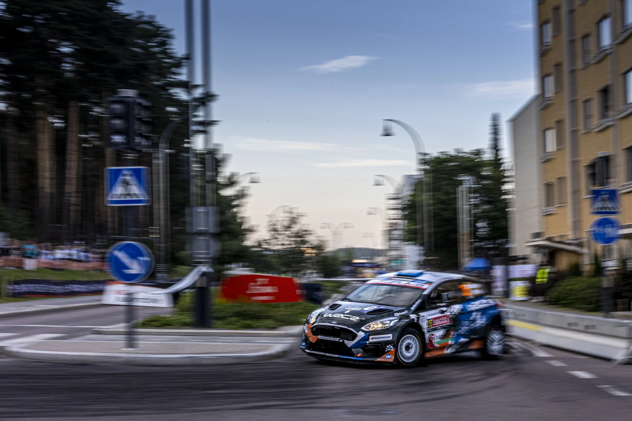 A racing car on a city street during a race, with blurred buildings and street signs in the background.