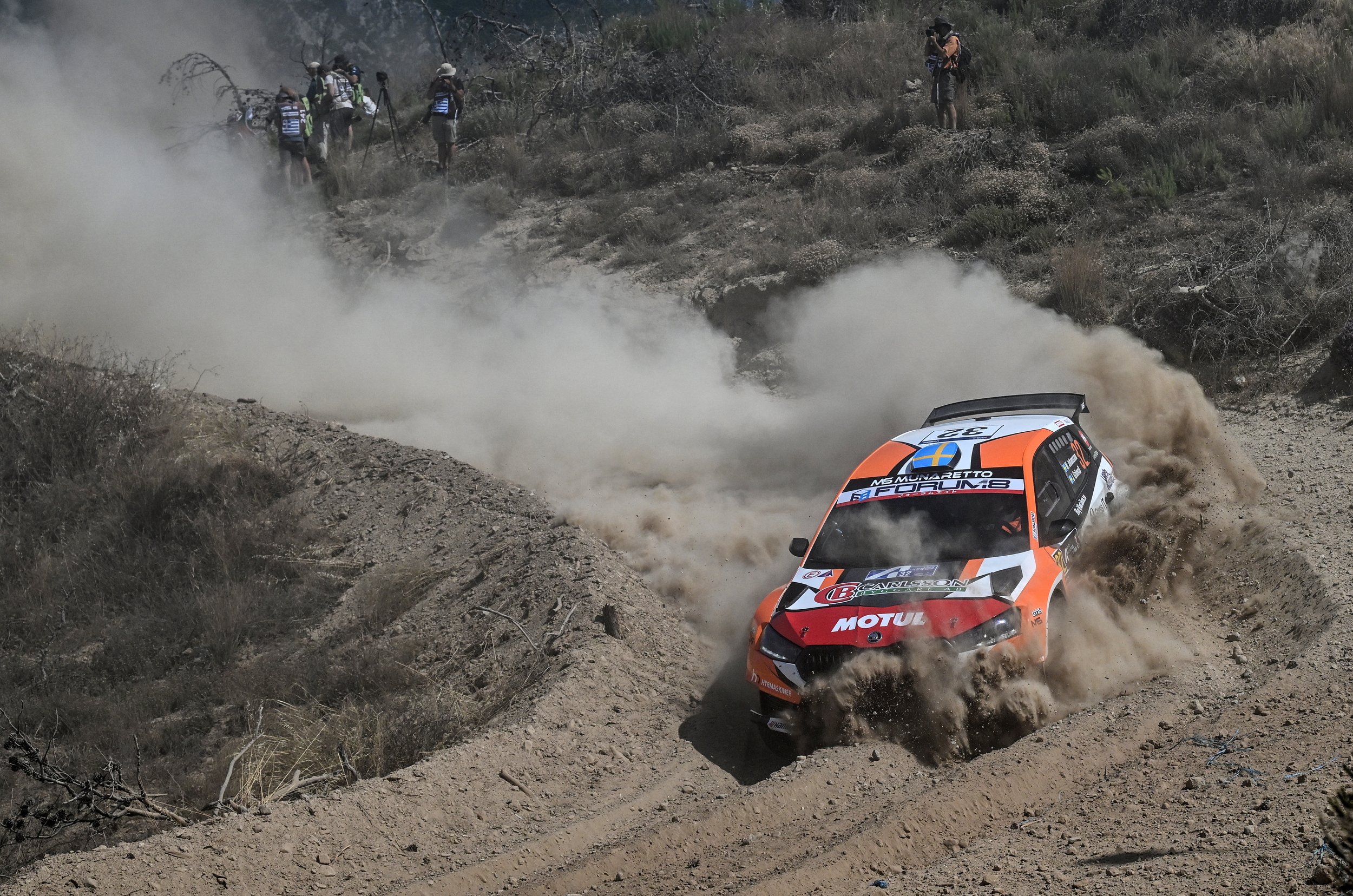 An orange and white rally car kicking up dust as it races on a dirt track, with several spectators and cameras on the hillside.