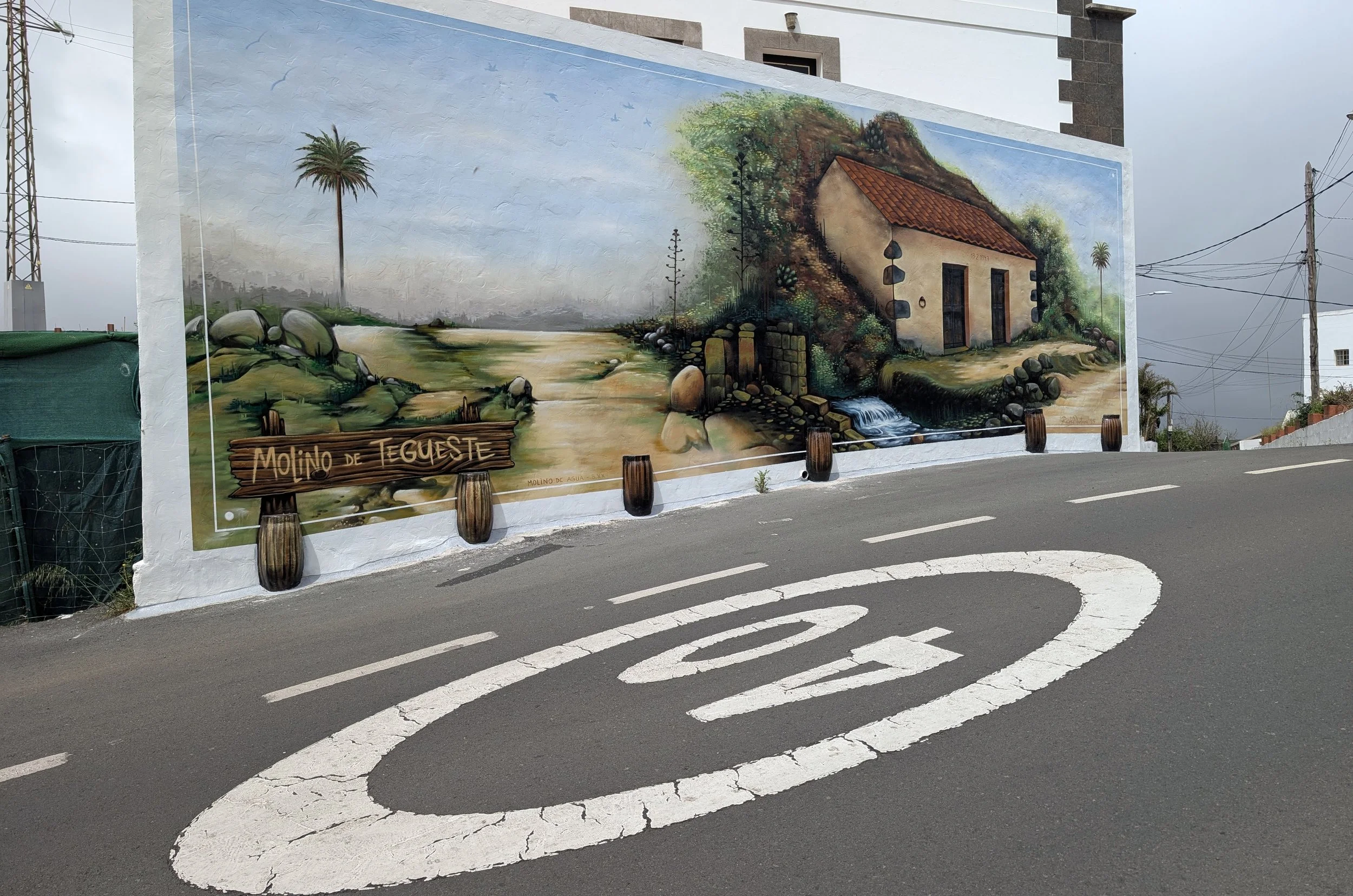 Colorful mural depicting a rustic house, a dirt path, lush greenery, trees, and a mountain landscape on a white wall next to a road with painted white lines and tire marks.