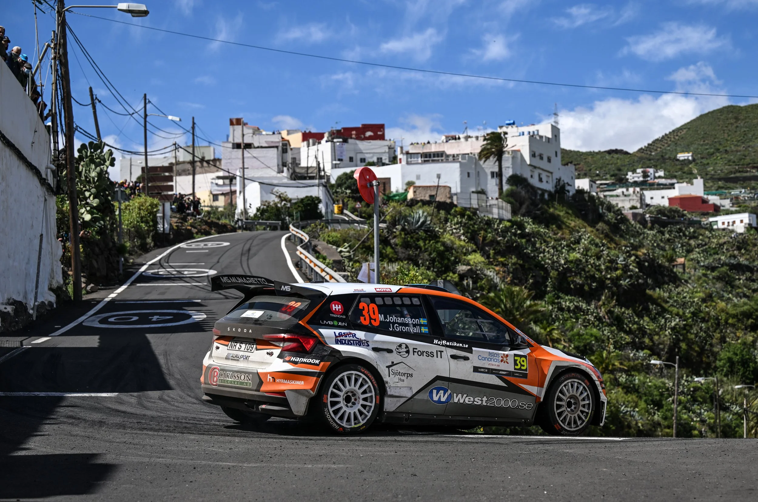A rally car on a winding mountain road, with hillside houses and bright blue sky in the background.