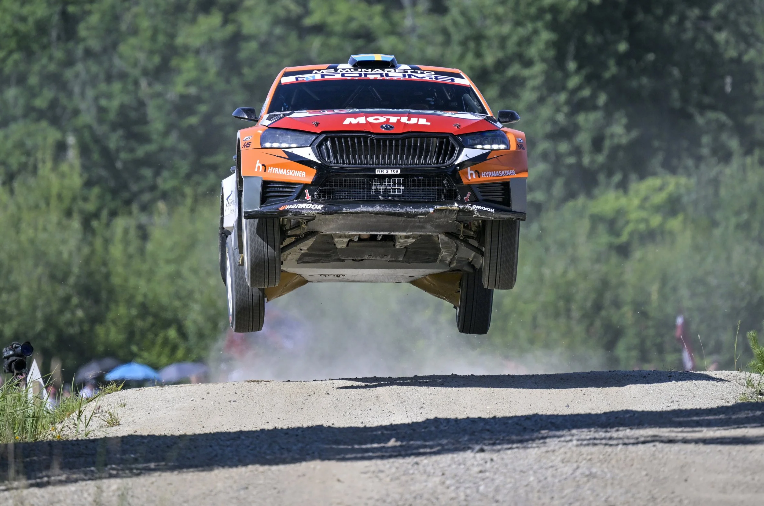 Race car airborne above dirt track during race with green forest background.
