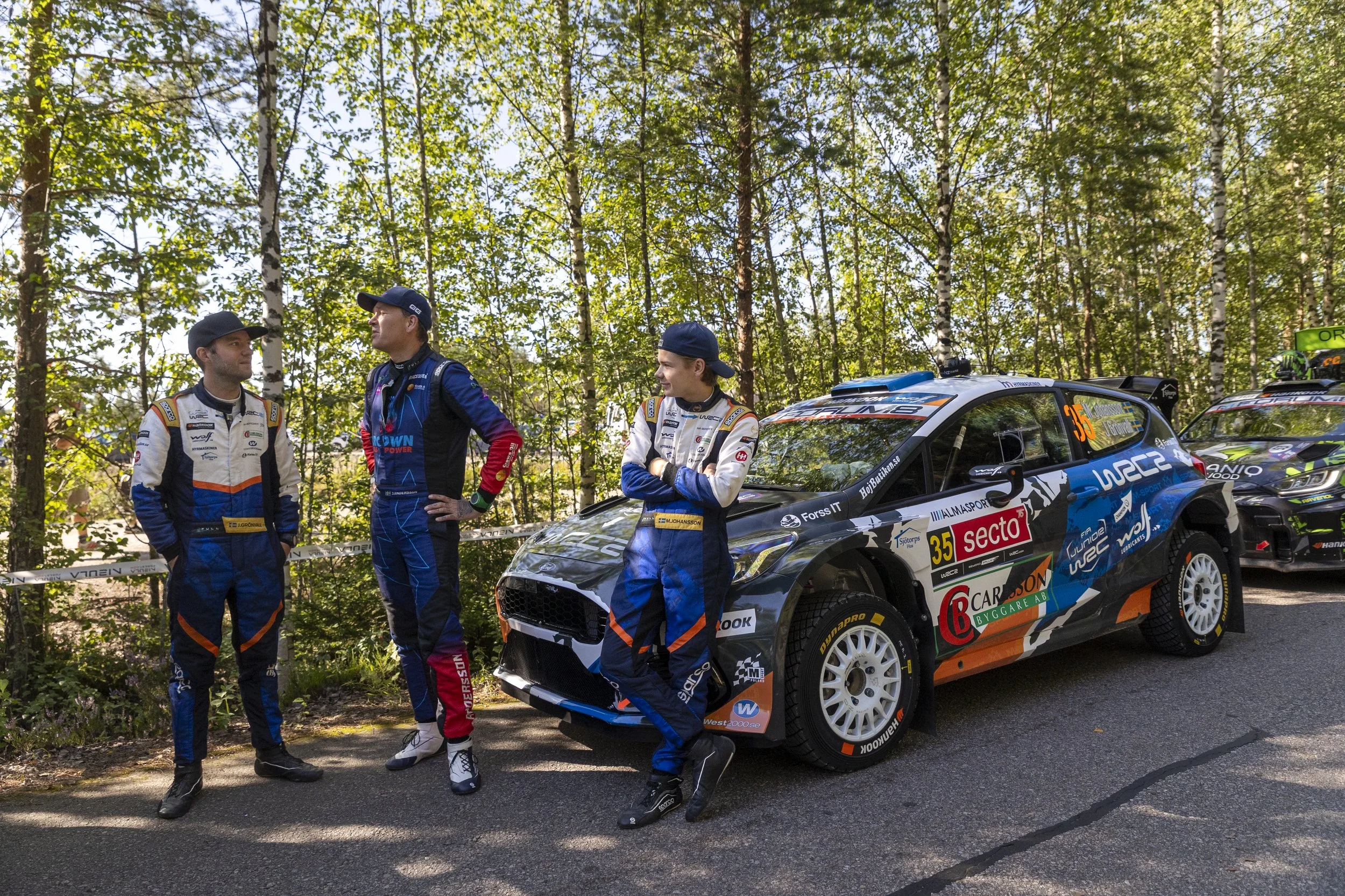 Three rally race drivers in racing suits and caps standing next to a rally car in a wooded area with trees and sunlight filtering through the leaves.