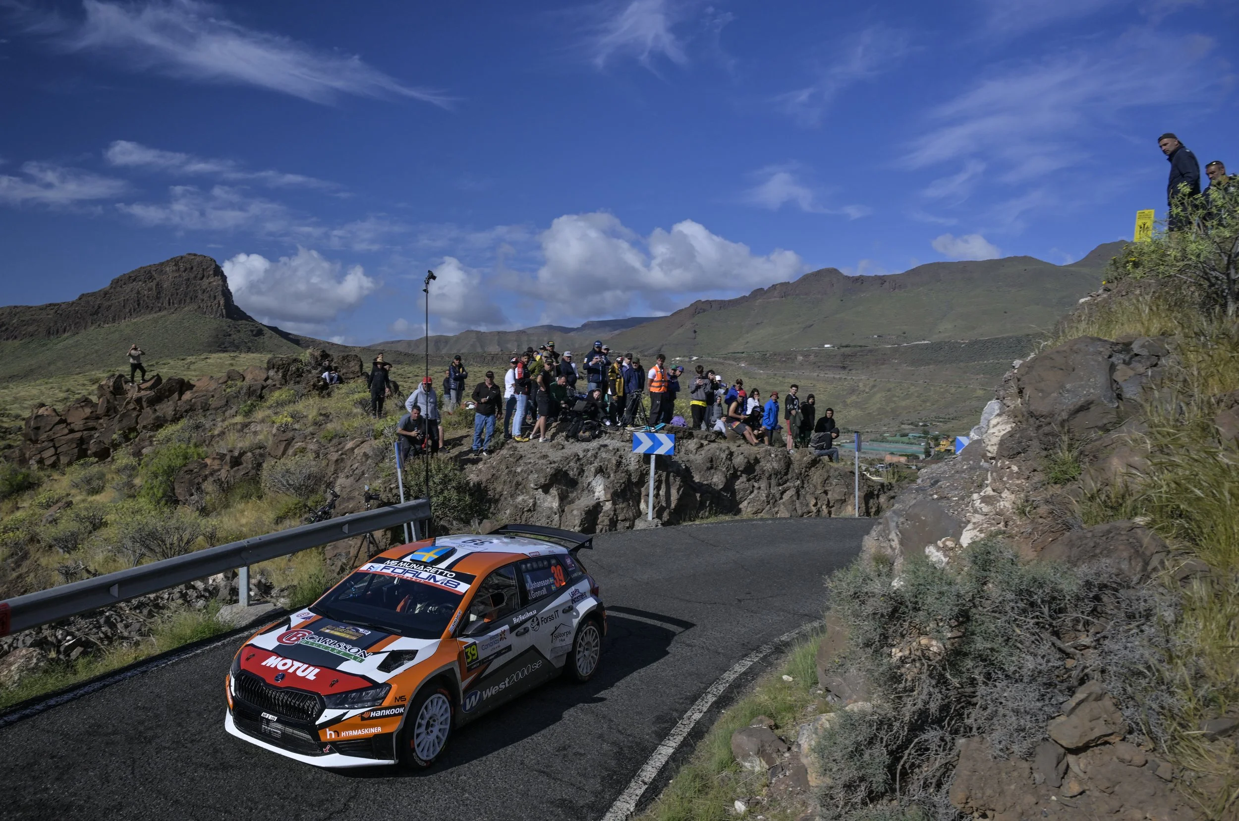 Rally car racing on a mountain road with spectators watching from the side, blue sky and clouds in the background.