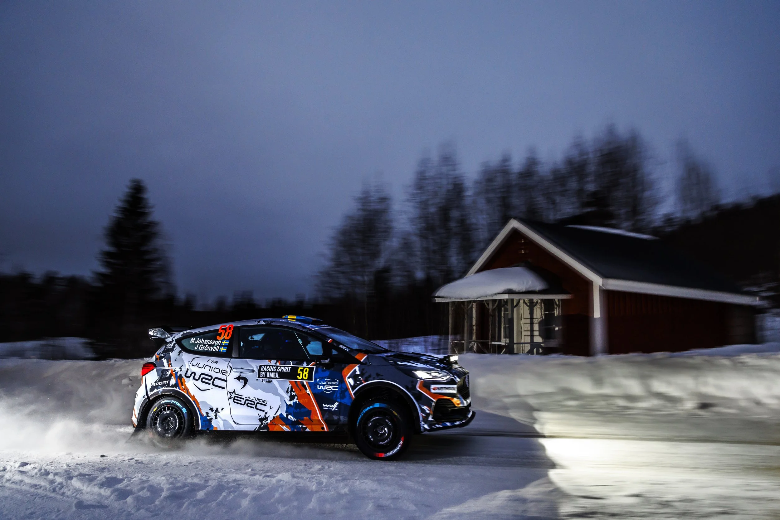 A white and black rally car with orange accents driving on snow during dusk or dawn, with a house and trees in the background.