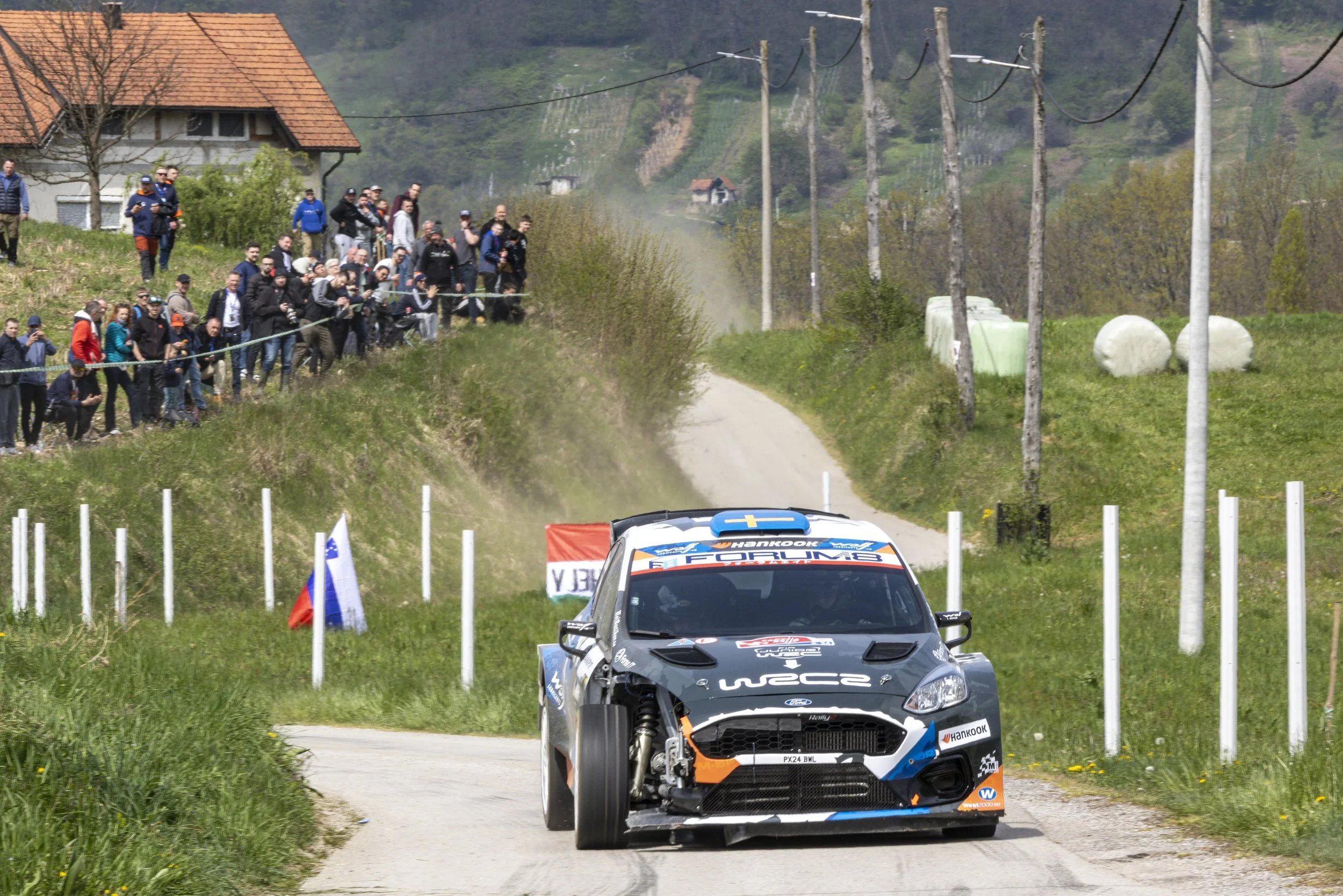 A rally car on a dirt road during a race, with spectators watching from the roadside, rural landscape, power lines, and hay bales in the background.