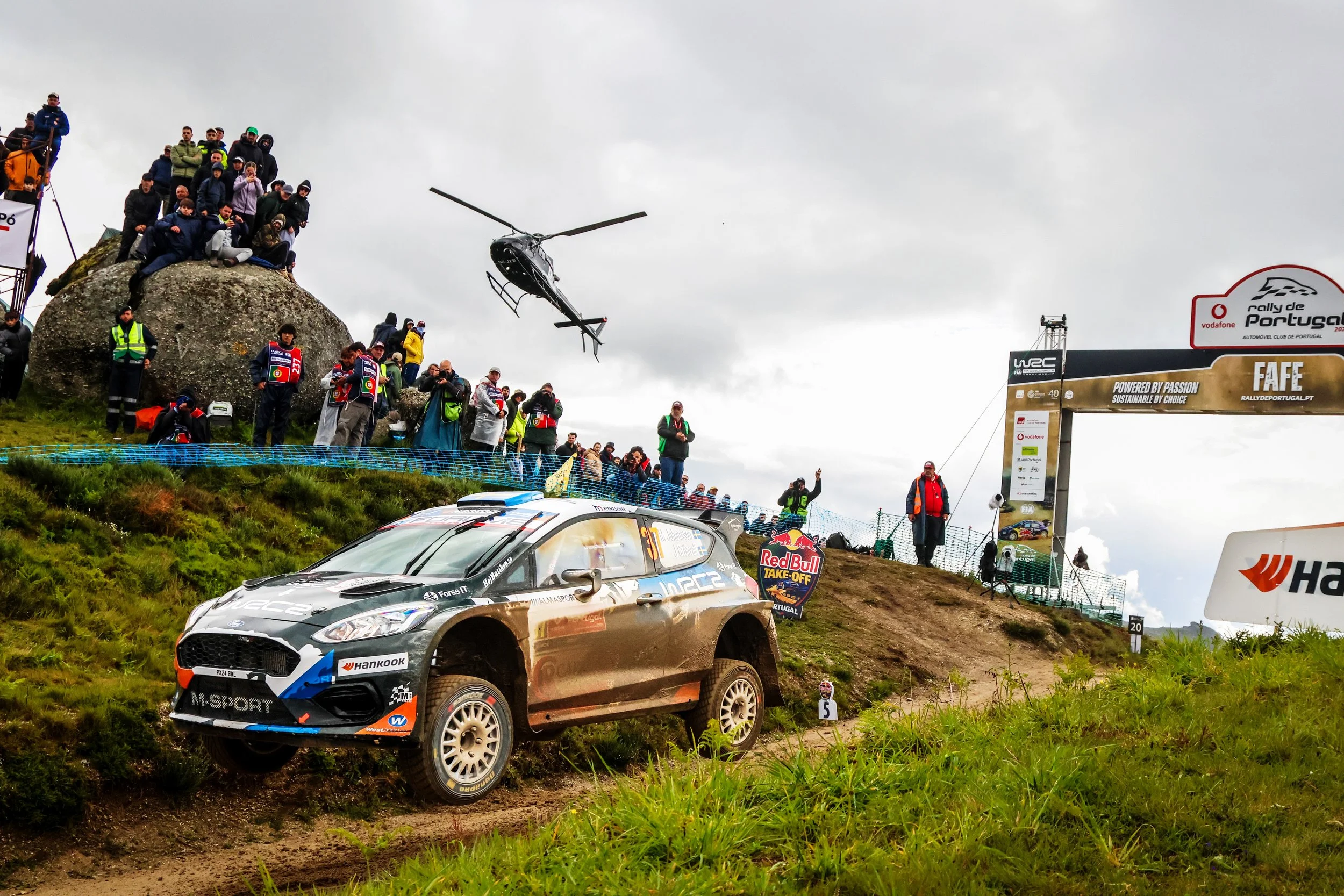 A rally car on a dirt track, with a helicopter flying overhead, and spectators and officials on the hillside.