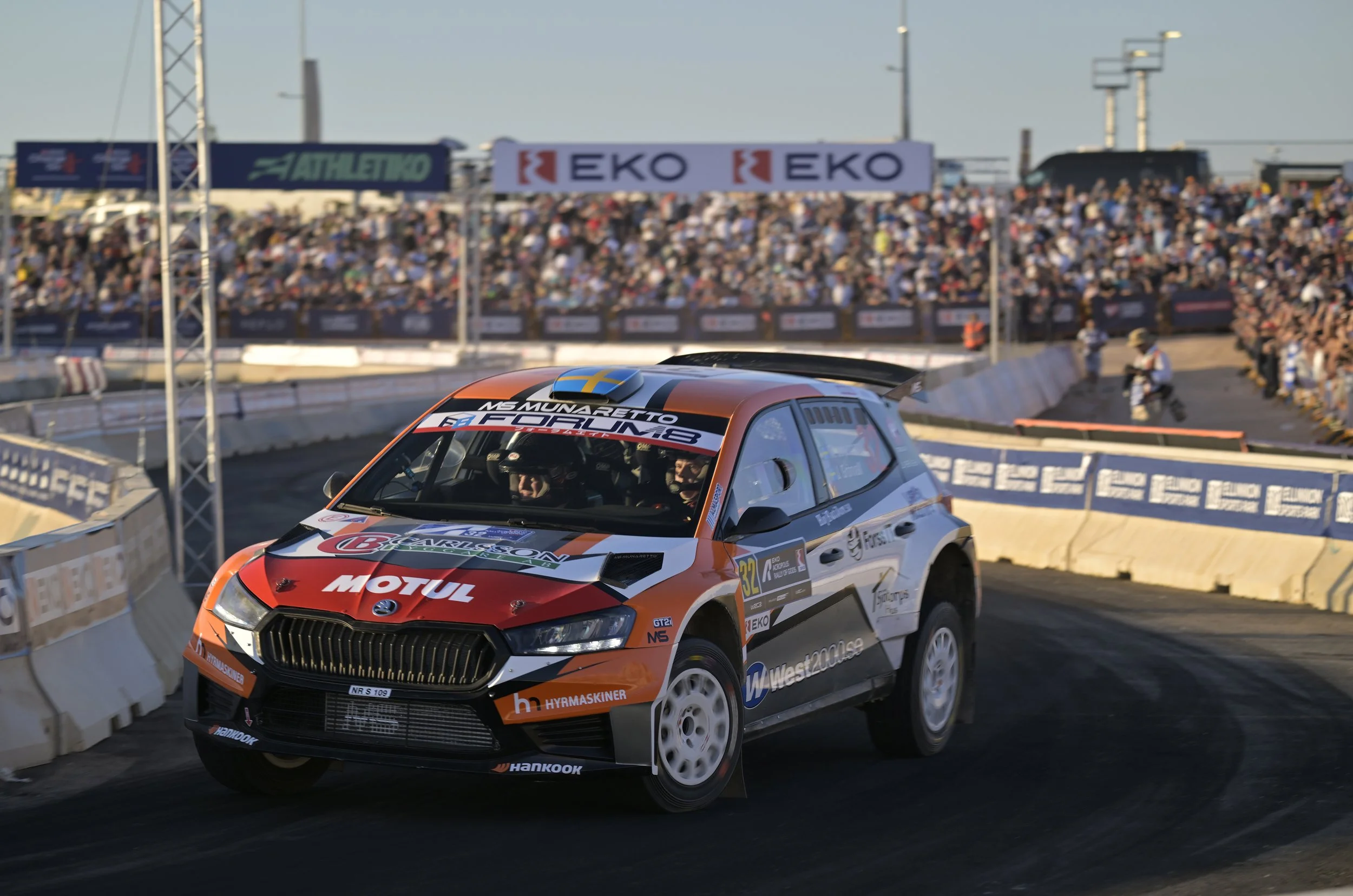 A rally car racing on a dirt track with a large crowd of spectators in the background, with banners and advertisements visible.