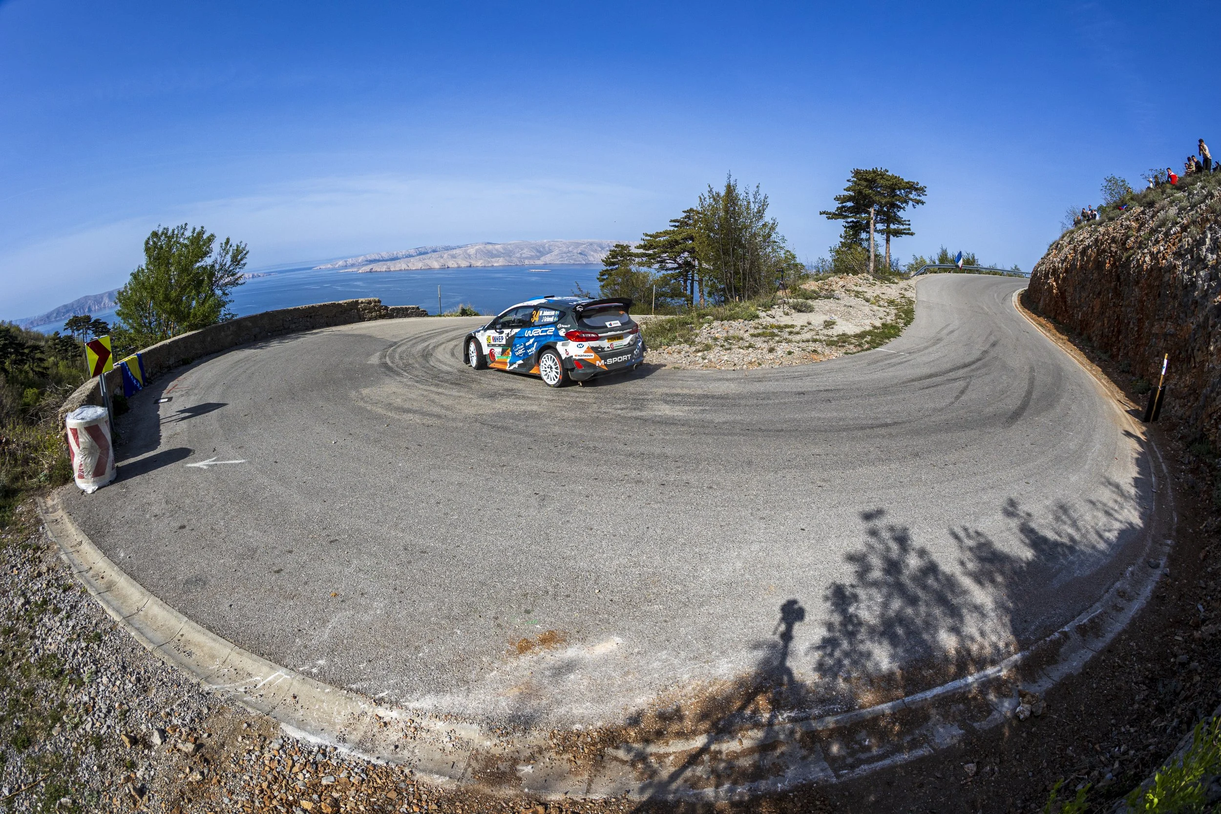 A rally car drives on a winding mountain road with cliffs on one side and scenic views of water and distant hills in the background. Spectators stand on the cliffs watching the race.