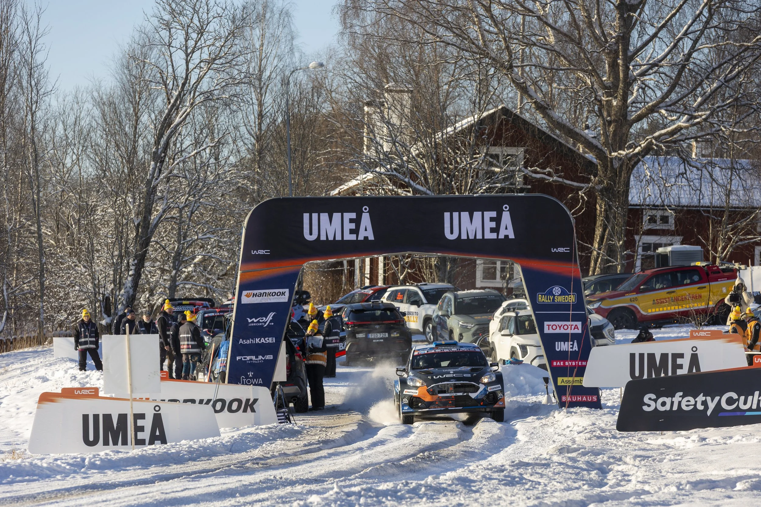 Winter rally event scene with cars, spectators, and banners in Umeå, Sweden, snowy landscape with leafless trees.