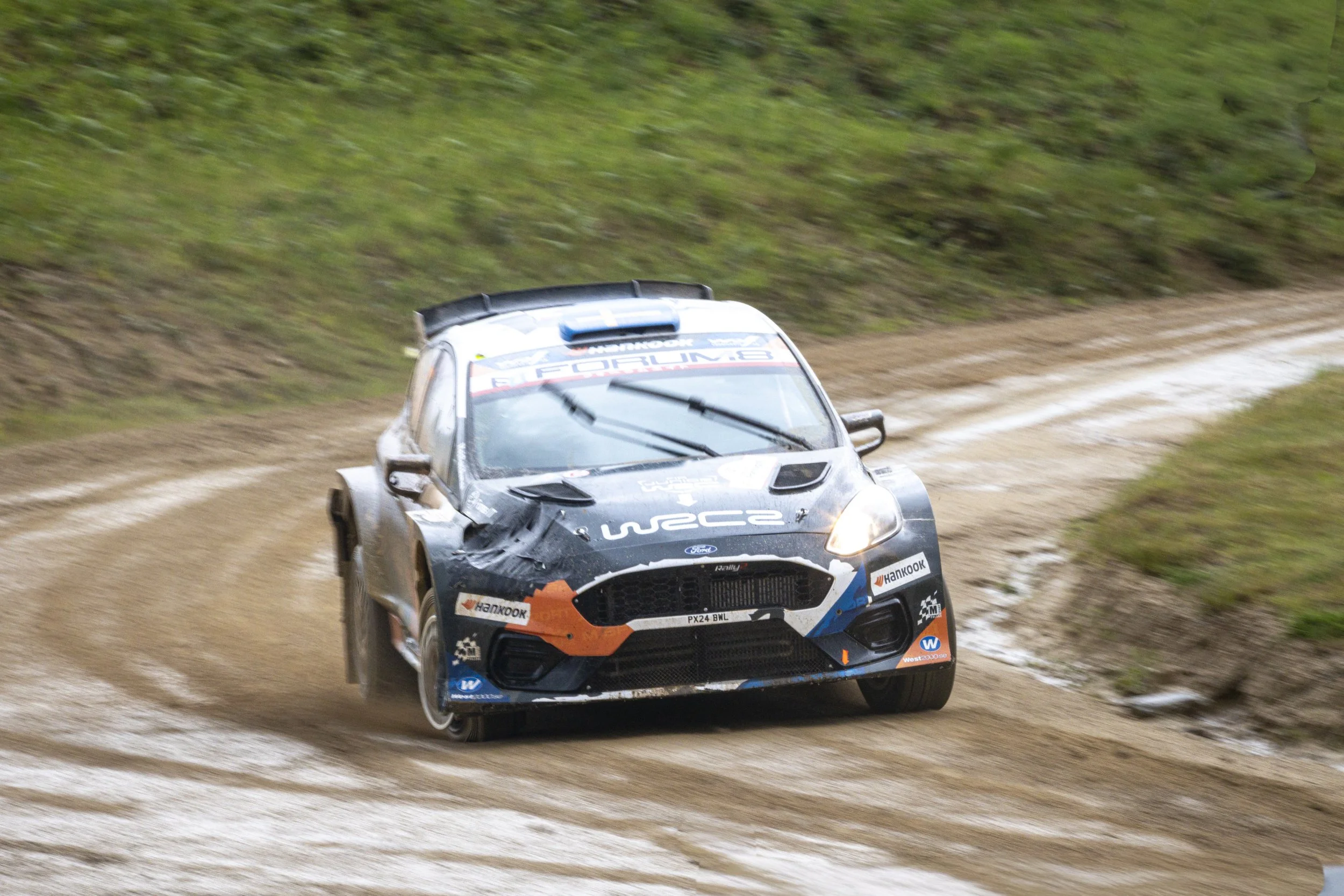 A rally car driving on a dirt road, kicking up dust in a grassy outdoor setting.